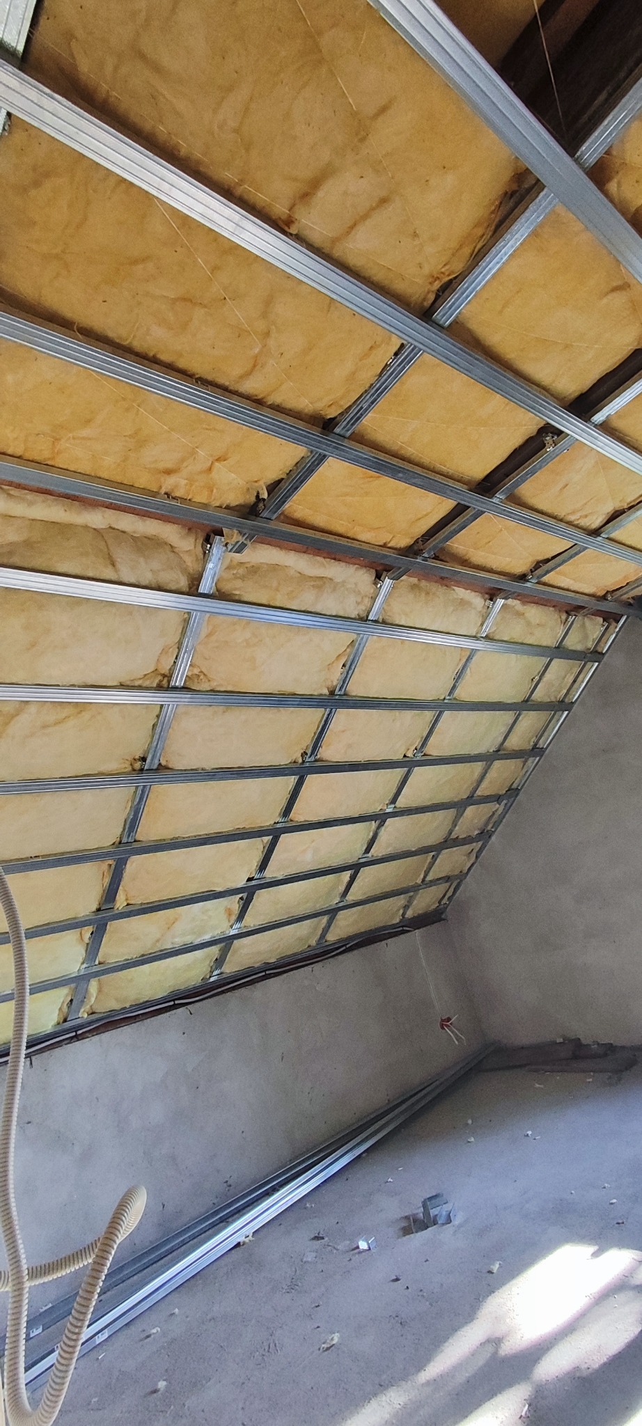 Poddasze w trakcie izolacji: wełna mineralna umieszczona w metalowym stelażu. Na podłodze leżą profile, widoczne kable elektryczne.