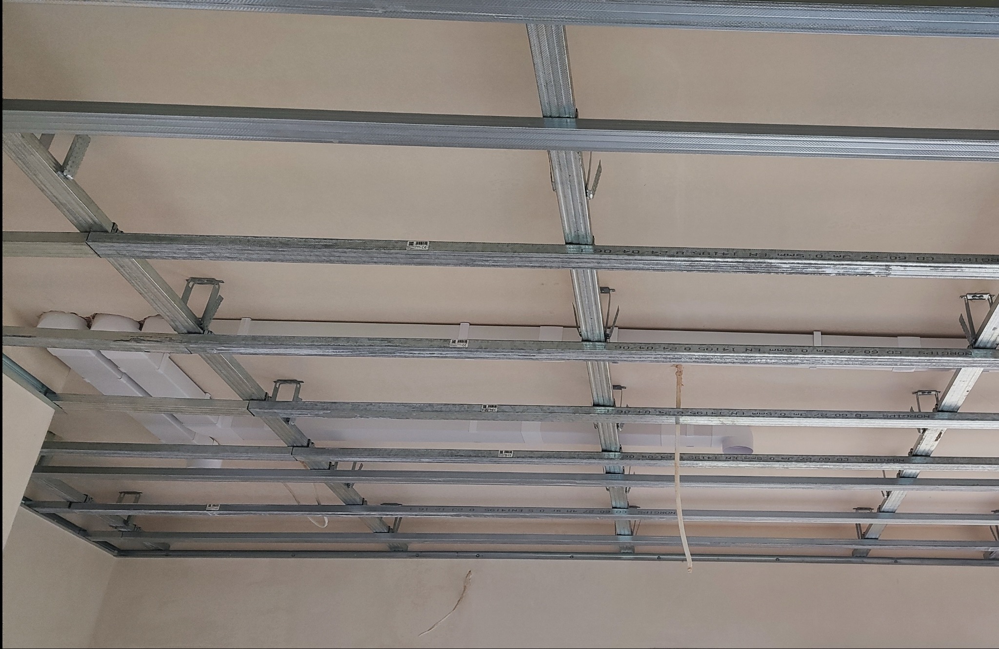 Stelaż metalowy sufitu podwieszanego z płyt kartonowo-gipsowych, widok od dołu, widoczne profile CD zamocowane do stropu za pomocą wieszaków ES, białe rury wentylacyjne w przestrzeni między sufitem...