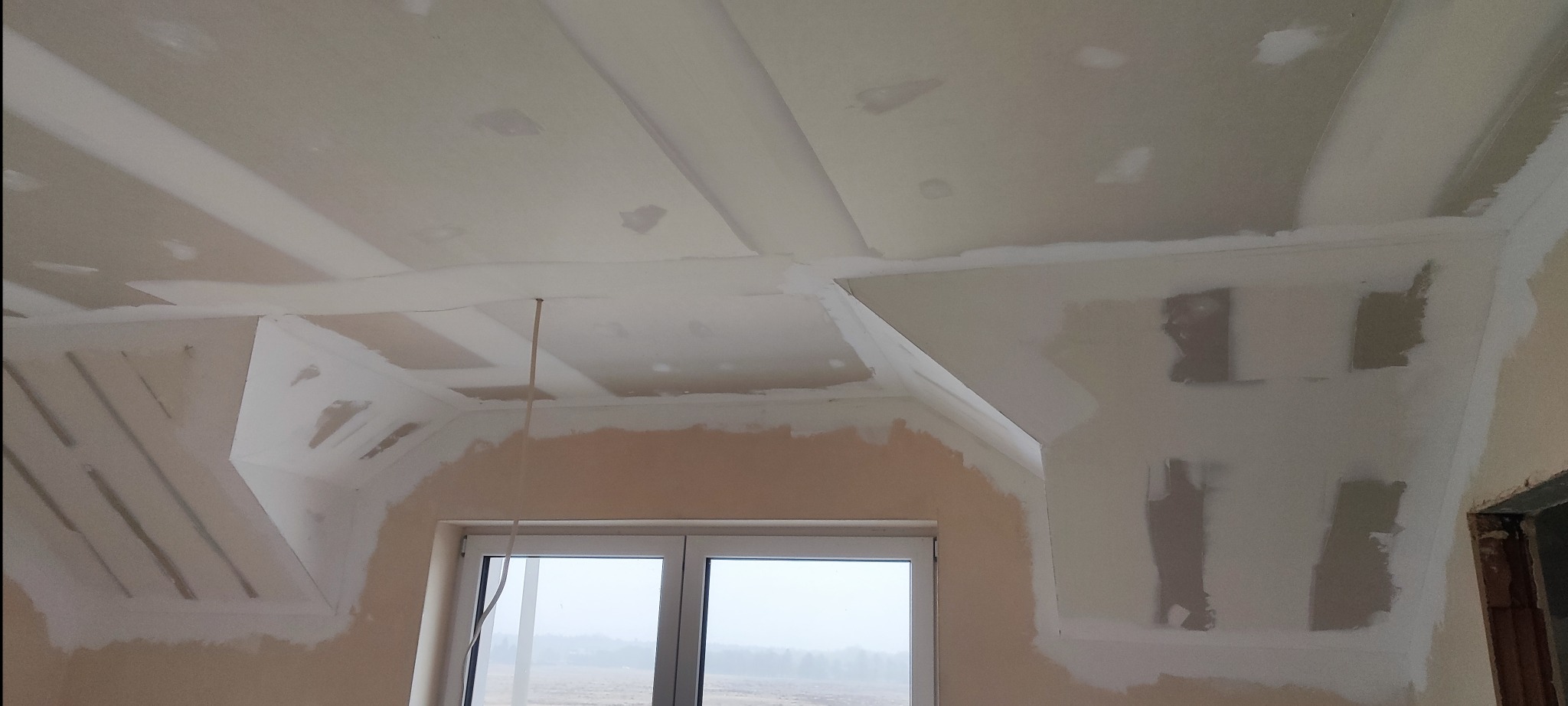 Poddasze z oknem dachowym w trakcie prac wykończeniowych, widoczne płyty kartonowo-gipsowe na skosach i suficie, wstępnie wyszpachlowane łączenia i fragmenty ścian, przewód elektryczny zwisający...