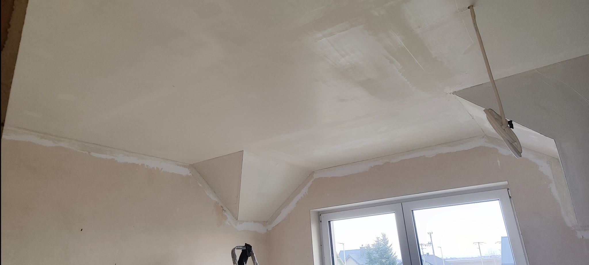Poddasze w trakcie wygładzania, widoczne świeżo nałożone warstwy gładzi na suficie i ścianach, narzędzie do rozprowadzania gładzi zawieszone u góry, okno w tle.