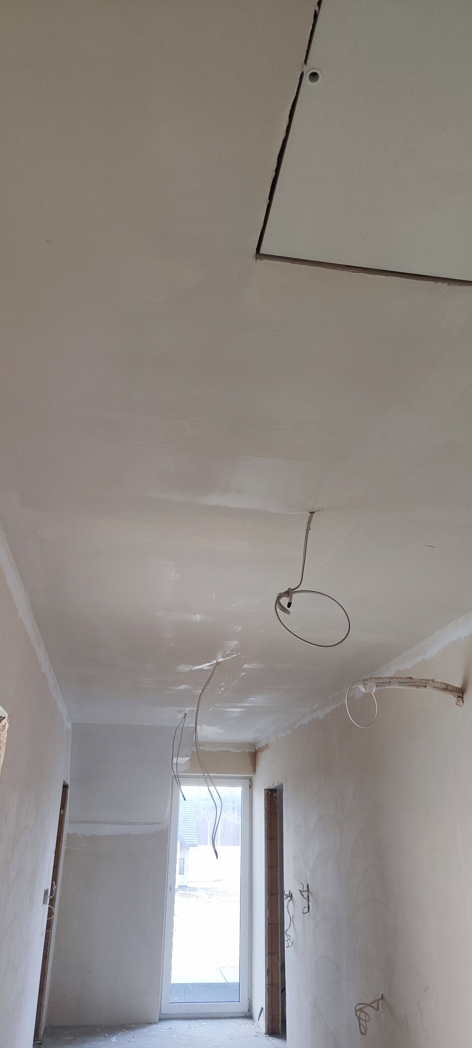 Wnętrze w trakcie prac wykończeniowych: sufit z widocznymi śladami szpachlowania, wystające przewody elektryczne i fragment płyty kartonowo-gipsowej na suficie, ściany przygotowane do malowania...