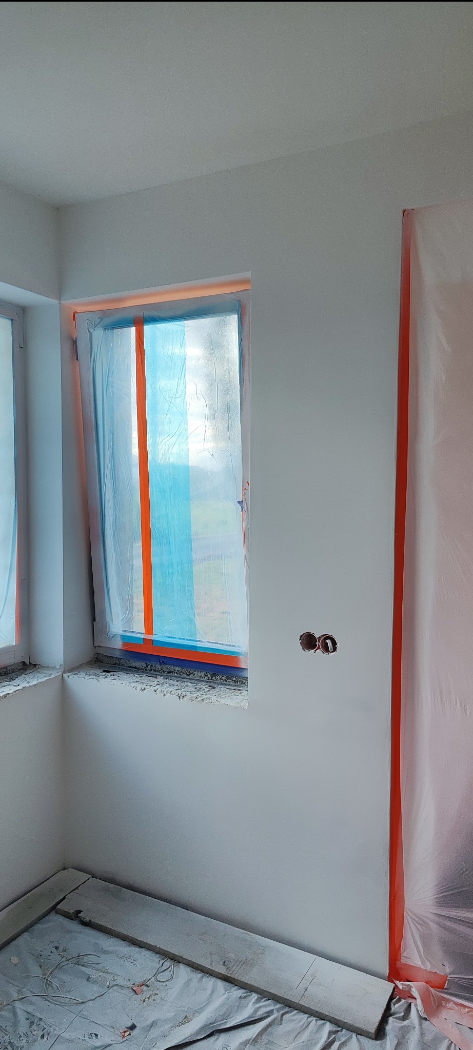 Przygotowanie do malowania ścian: zabezpieczone okna folią i taśmą, zakryta podłoga, świeżo pomalowane ściany, puszki elektryczne bez osprzętu.