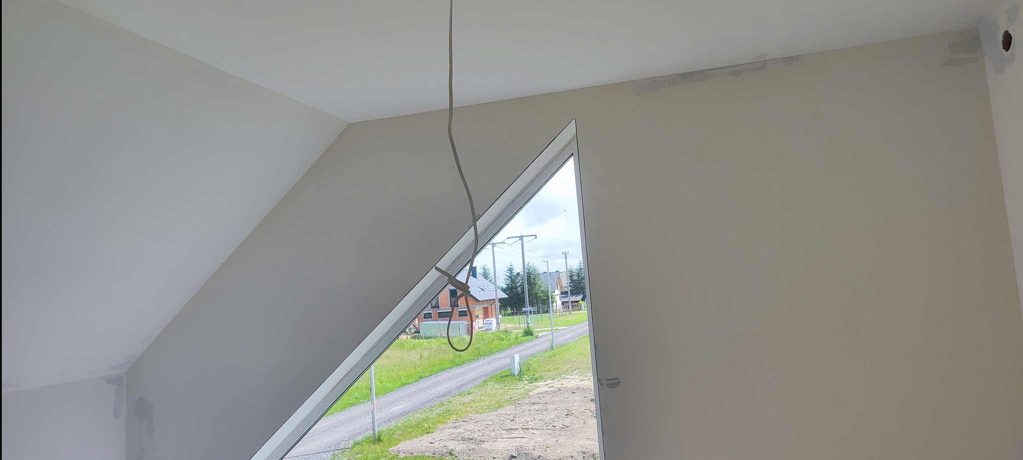 Poddasze po gładziowaniu, widoczne niedoskonałości przy suficie i okablowanie zwisające z sufitu. Trójkątne okno z widokiem na zewnątrz.