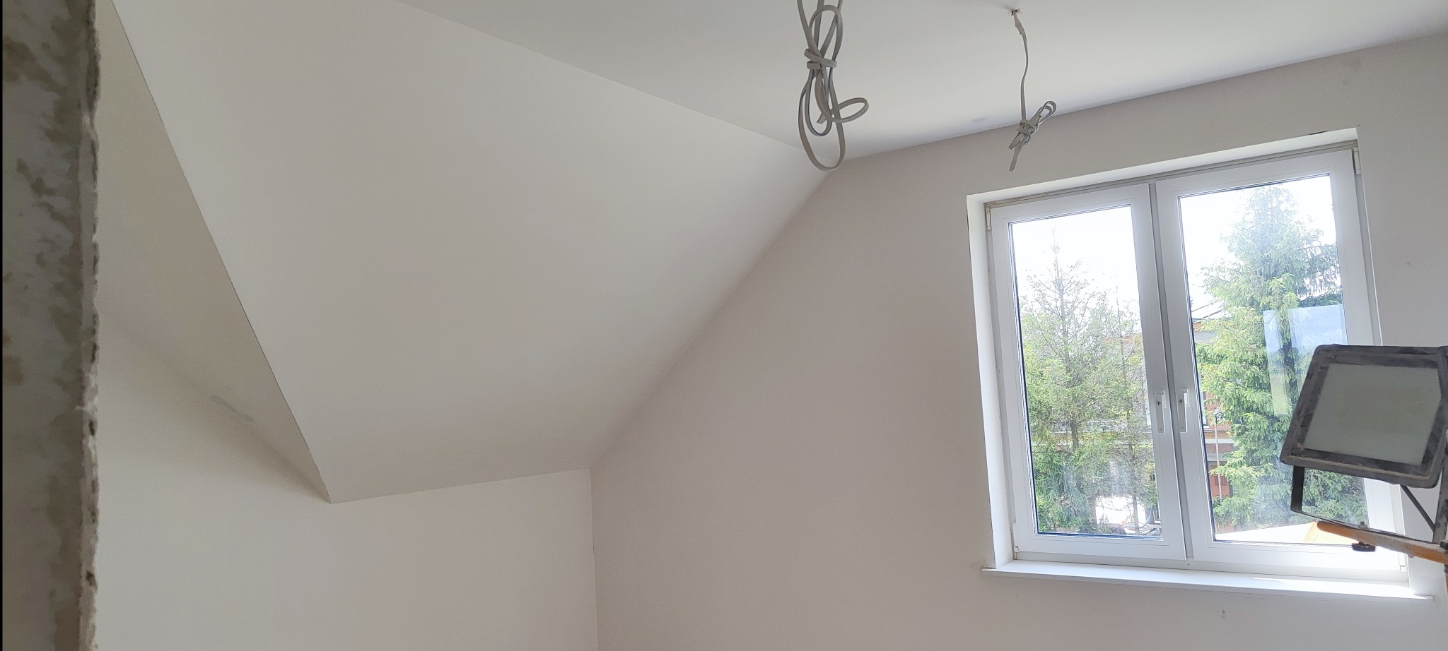 Poddasze po gładzi, ściany i skos sufitu pomalowane na biało. Widoczne kable oświetleniowe i okno z widokiem na zieleń. W rogu lampa doświetlająca.