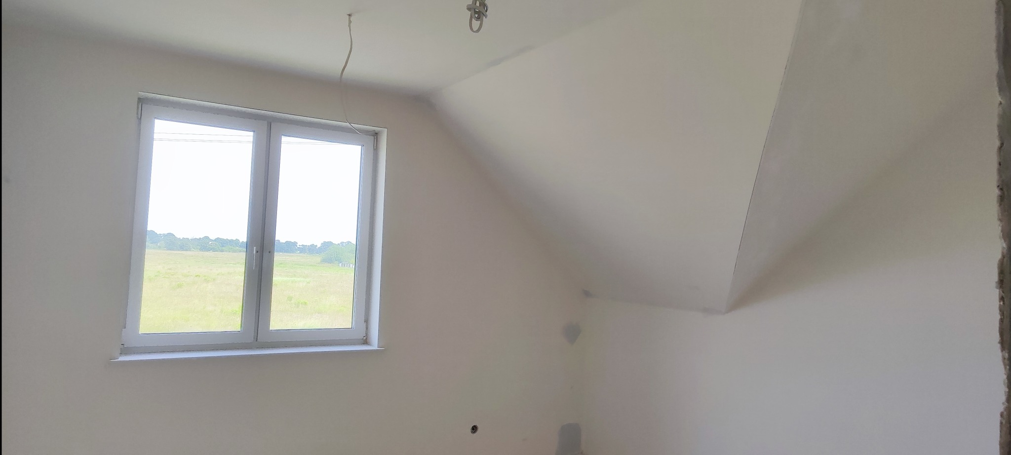Poddasze po szpachlowaniu, jasne ściany, okno z widokiem na łąkę. Widoczne ślady po szpachlowaniu na ścianach i suficie. Przygotowanie do malowania.