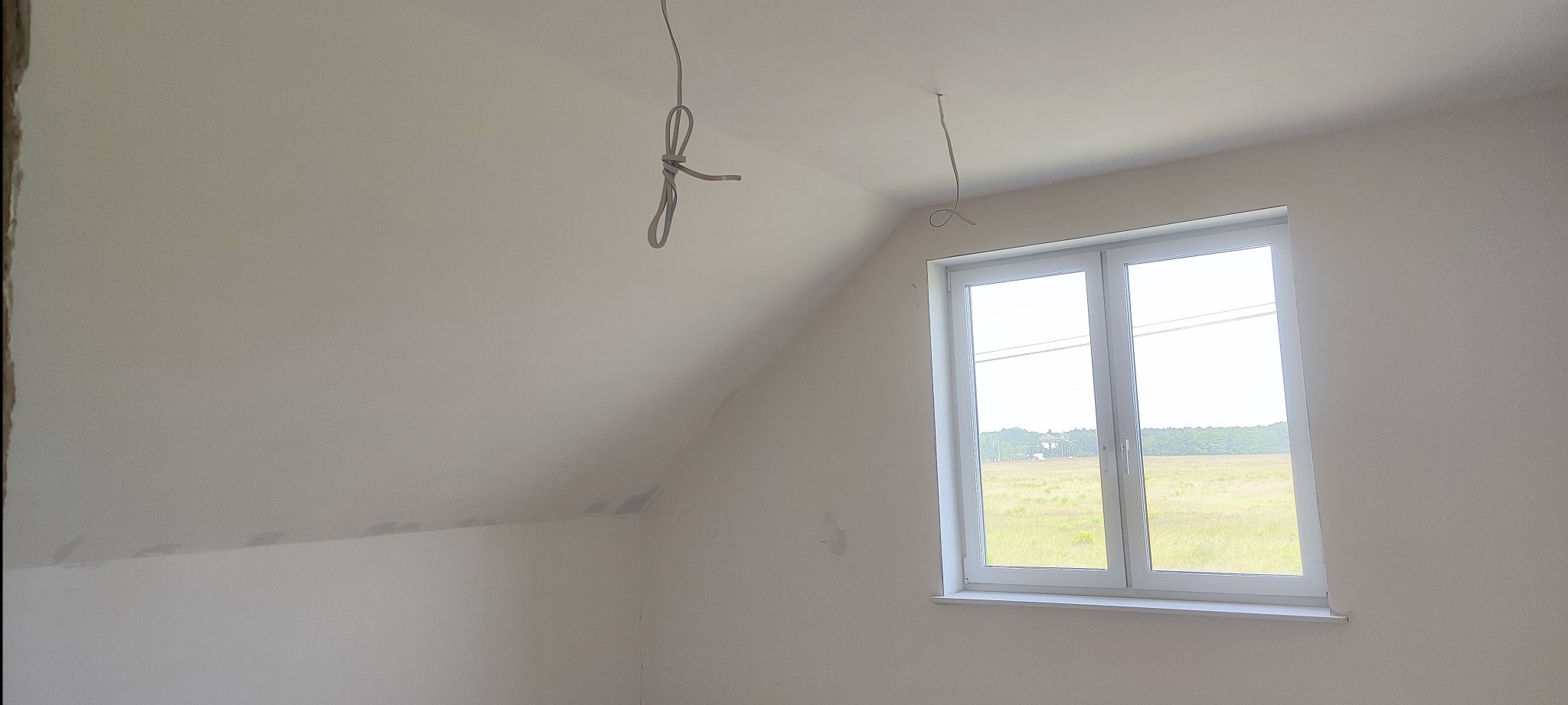 Poddasze po szpachlowaniu, z widocznymi przewodami elektrycznymi i oknem z widokiem na łąkę. Ściany przygotowane do malowania.