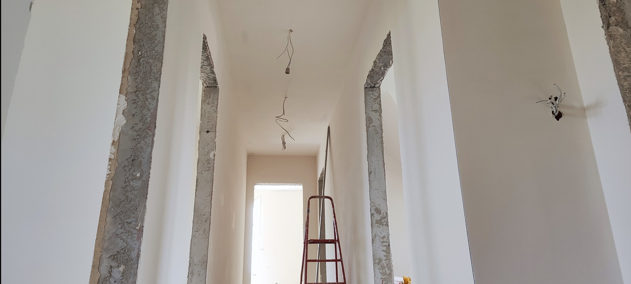 Korytarz w trakcie remontu: ściany po szpachlowaniu, widoczne kable oświetleniowe, drabina. Przygotowanie do malowania ścian i sufitu.