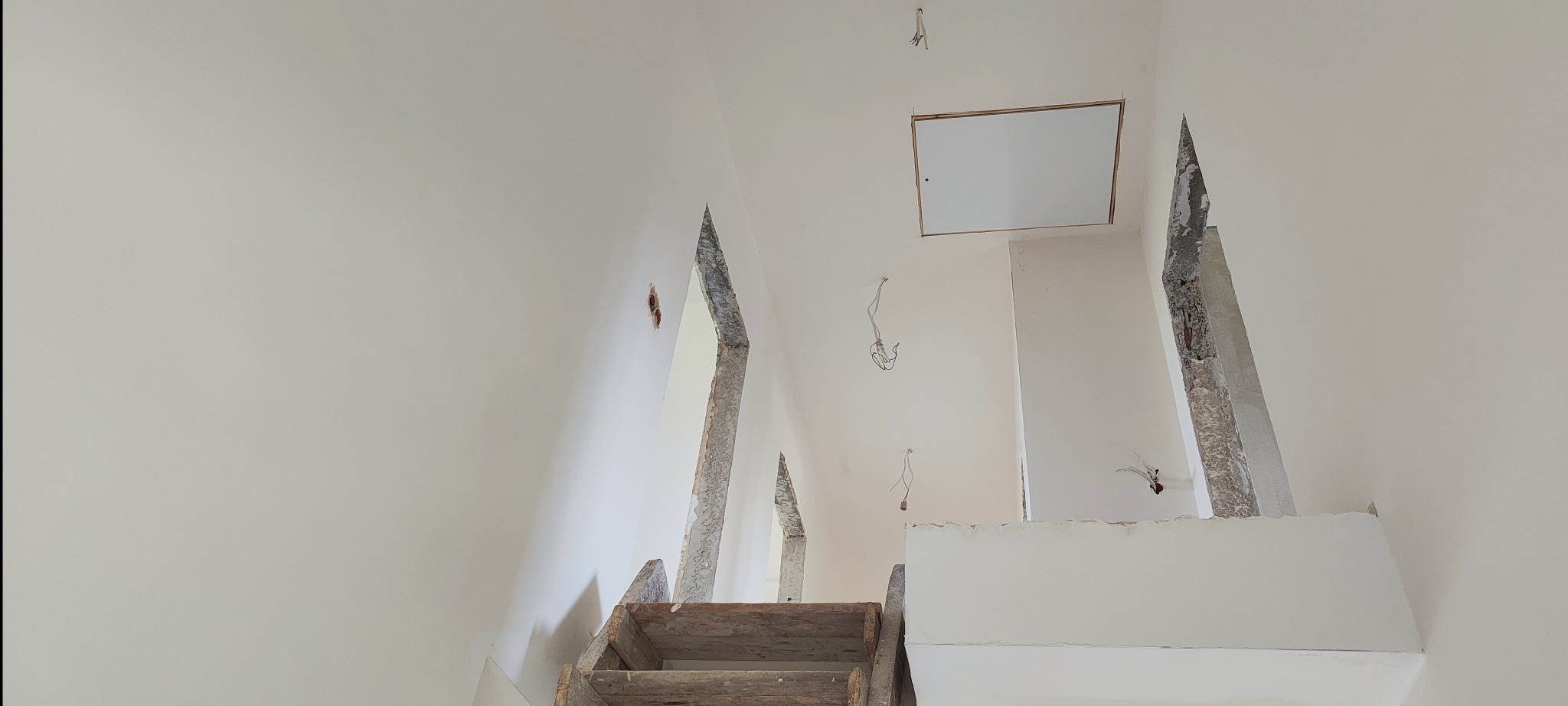 Białe, świeżo pomalowane wnętrze z drewnianymi schodami i otworami na kable. Widoczne elementy remontu, sufit z klapą rewizyjną.