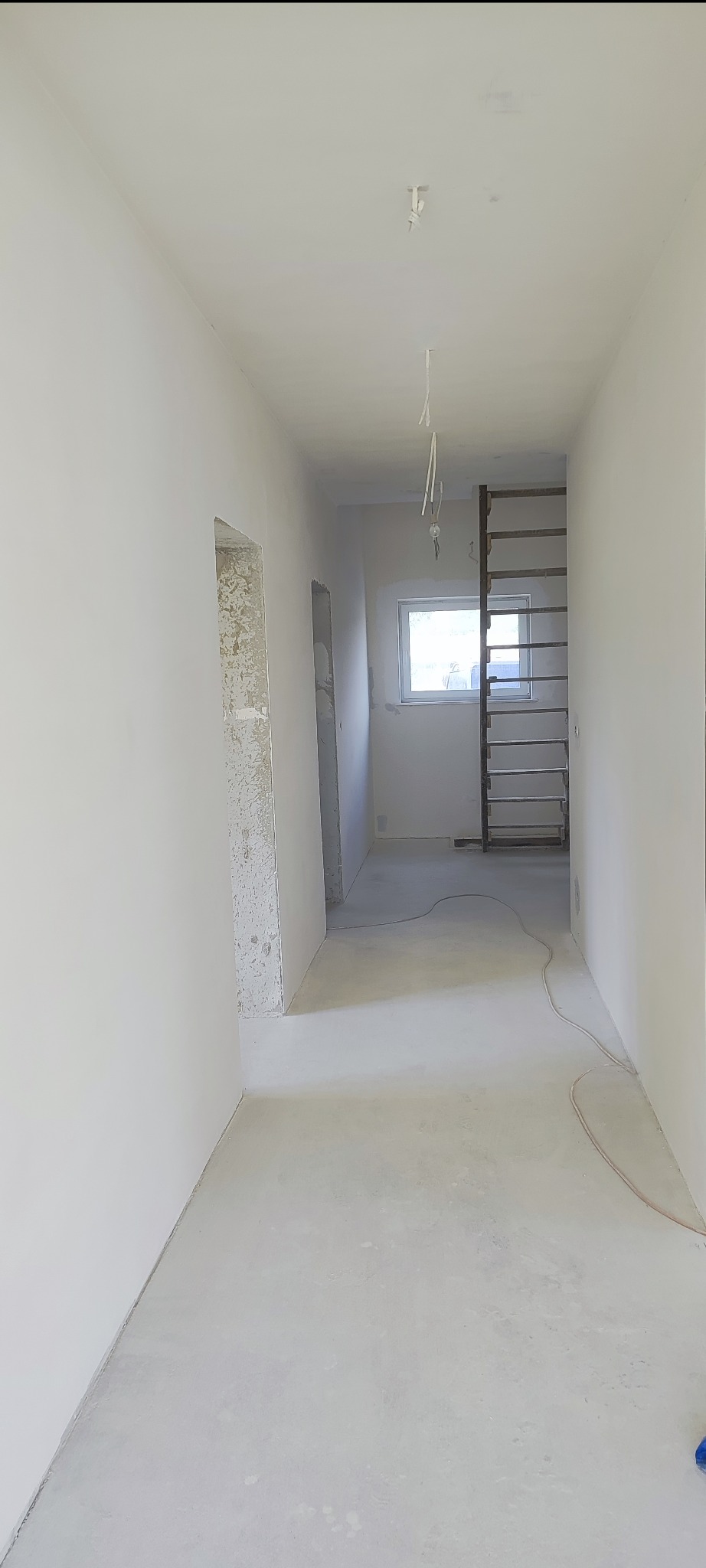 Wnętrze korytarza w trakcie prac wykończeniowych, widoczne gładkie, białe ściany, surowa podłoga i drabina oparta o ścianę przy oknie. Częściowo wykończone ościeża.