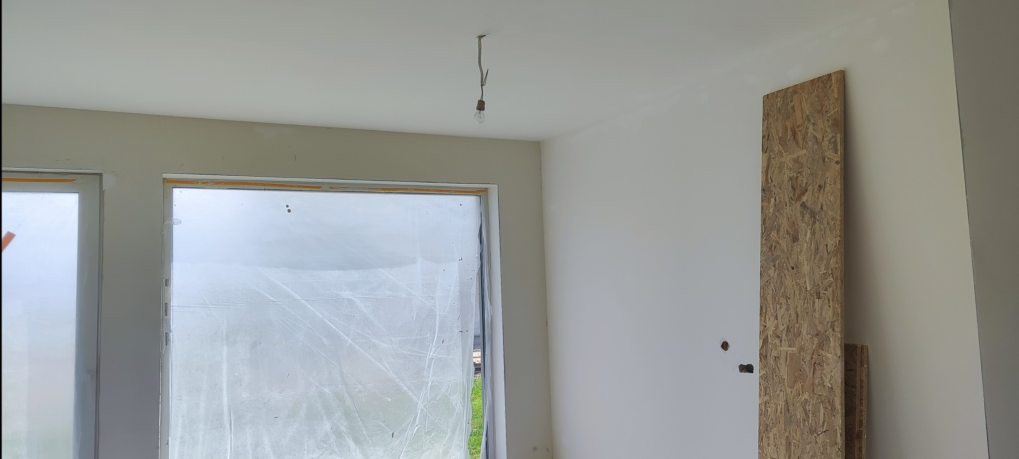 Wnętrze pokoju w trakcie prac wykończeniowych, z osłoniętymi oknami, gładkimi ścianami, wiszącą żarówką i płytą OSB opartą o ścianę.