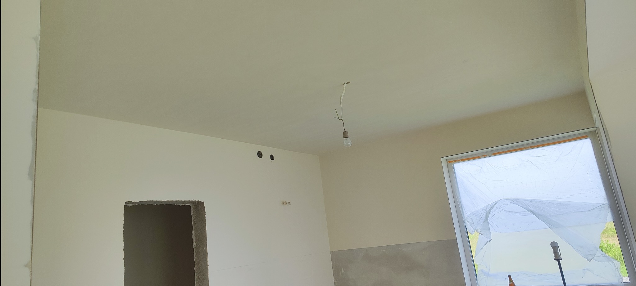Wnętrze po szpachlowaniu: gładkie ściany w jasnym odcieniu, widoczne otwory na instalacje, okno zabezpieczone folią. Żarówka na kablu zwisa z sufitu.