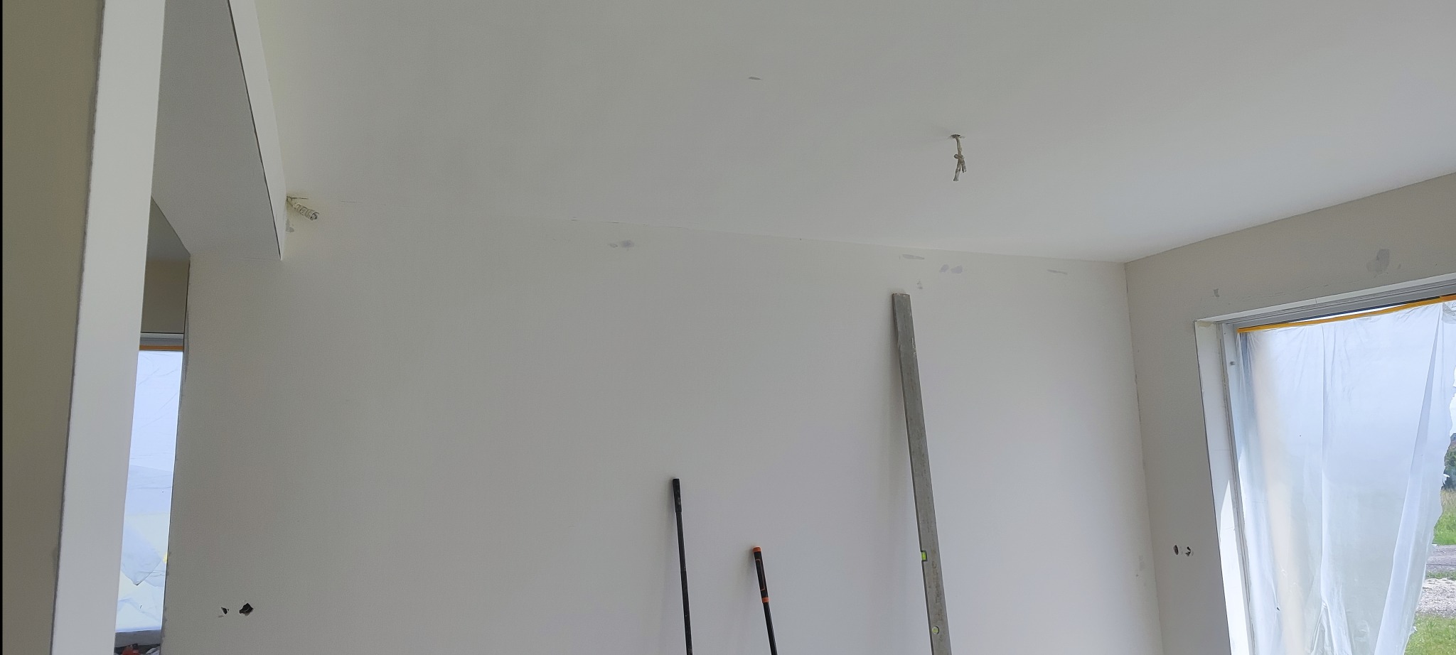 Wnętrze w trakcie wykańczania: białe ściany po szpachlowaniu, widoczne narzędzia (poziomica, kije), okno zabezpieczone folią malarską. Przygotowanie do malowania.