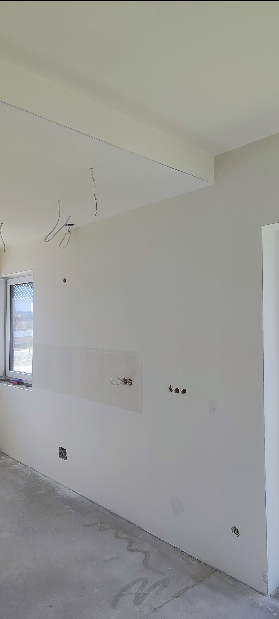 Wnętrze w trakcie remontu: gładkie, białe ściany z widocznymi przewodami elektrycznymi, przygotowane do dalszych prac wykończeniowych. Na podłodze ślady po pracach budowlanych.