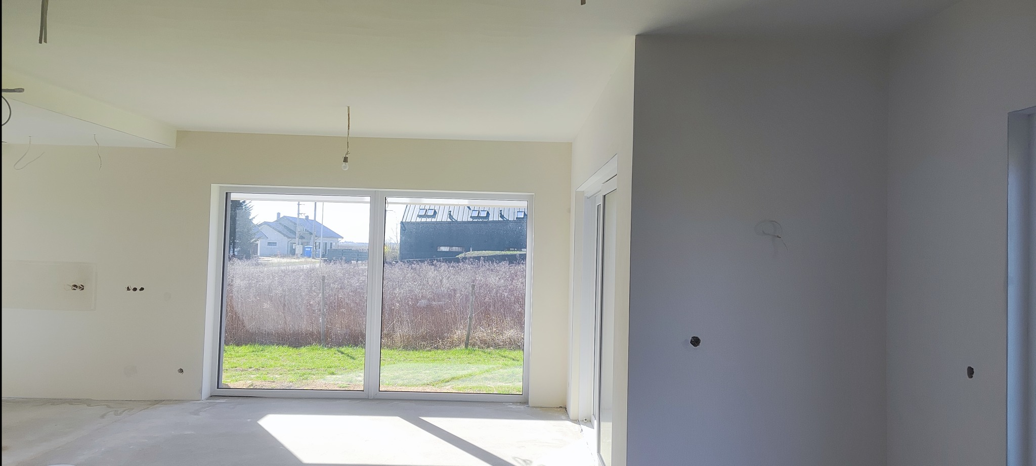 Jasne, przestronne wnętrze w trakcie wykańczania: gładkie ściany w odcieniach bieli i beżu, duże okno z widokiem na zieleń, surowy beton na podłodze. Widoczne instalacje elektryczne.