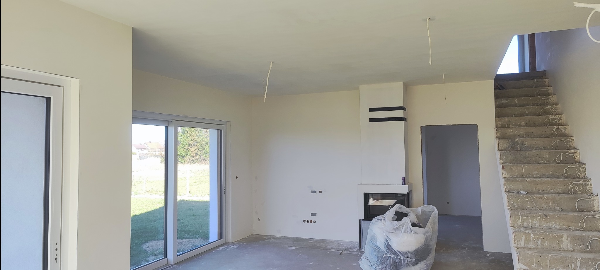 Wnętrze domu w trakcie wykańczania: świeżo pomalowane ściany w odcieniach bieli, surowe schody betonowe i kominek z czarnymi elementami dekoracyjnymi.