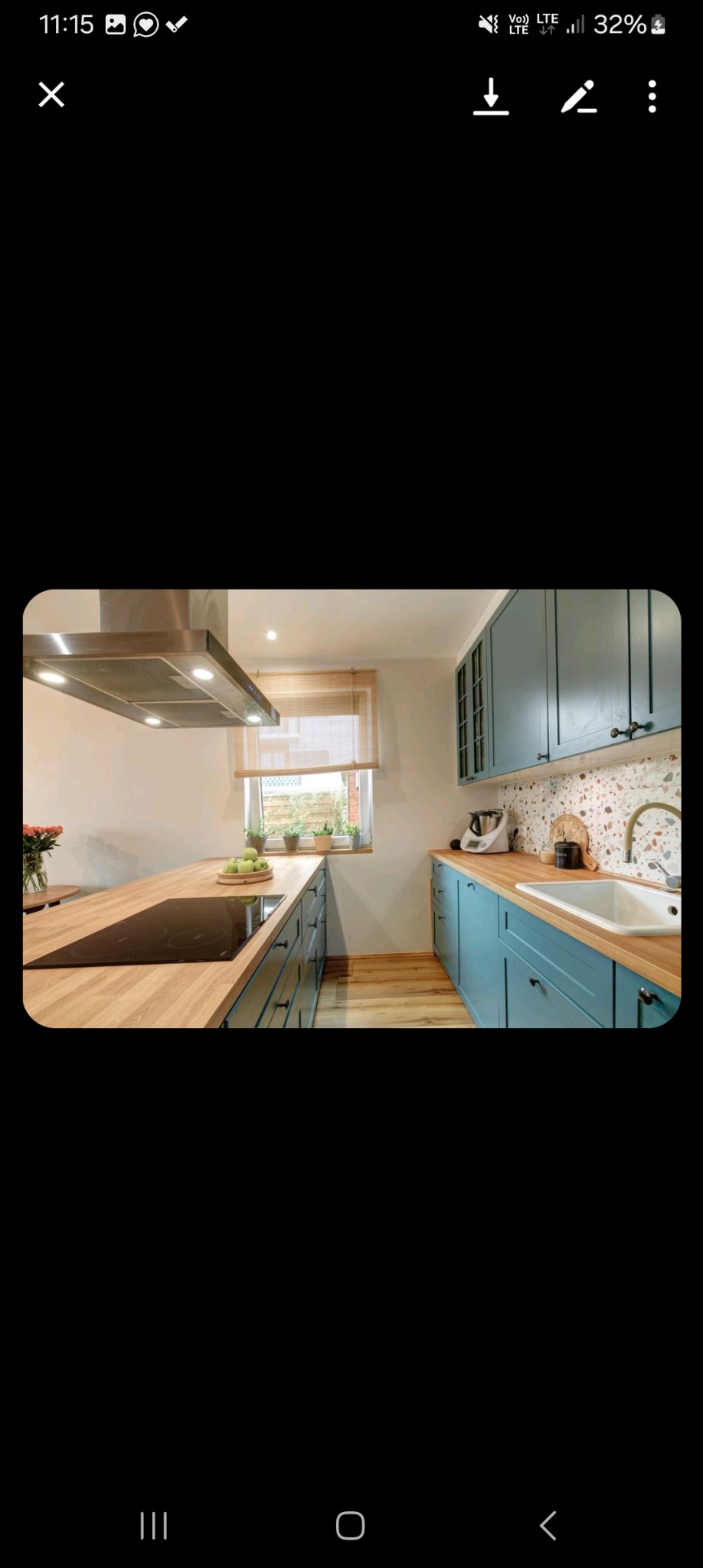 Turkusowa kuchnia z drewnianym blatem, płytą indukcyjną i okapem, widok na okno z bambusową roletą i roślinami.