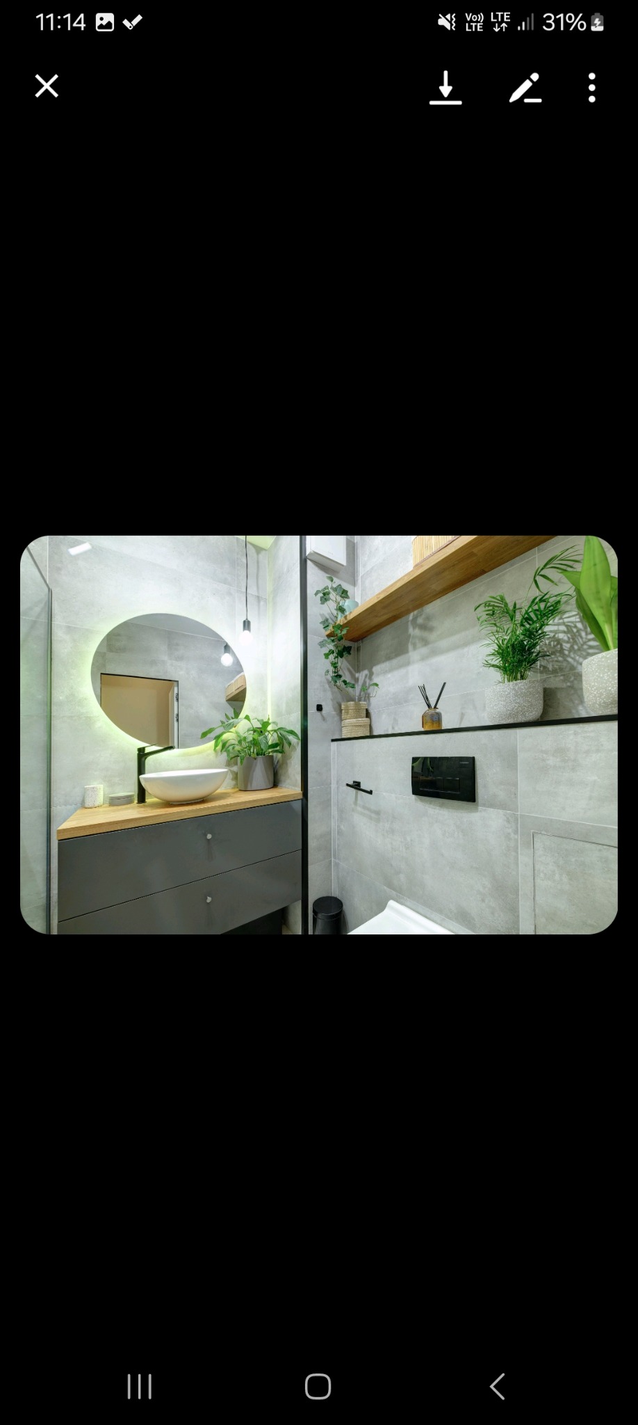 Nowoczesna łazienka z szarą glazurą, okrągłym lustrem z podświetleniem LED, drewnianym blatem pod umywalką i zielonymi roślinami doniczkowymi.