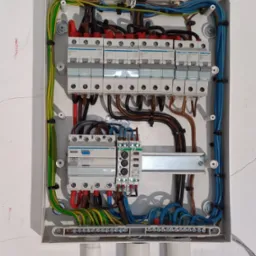 Otwarta skrzynka rozdzielcza z zainstalowanymi bezpiecznikami, przewodami w różnych kolorach (niebieski, żółto-zielony, brązowy, czerwony) i elementami montażowymi, widoczne białe rury instalacyjne...