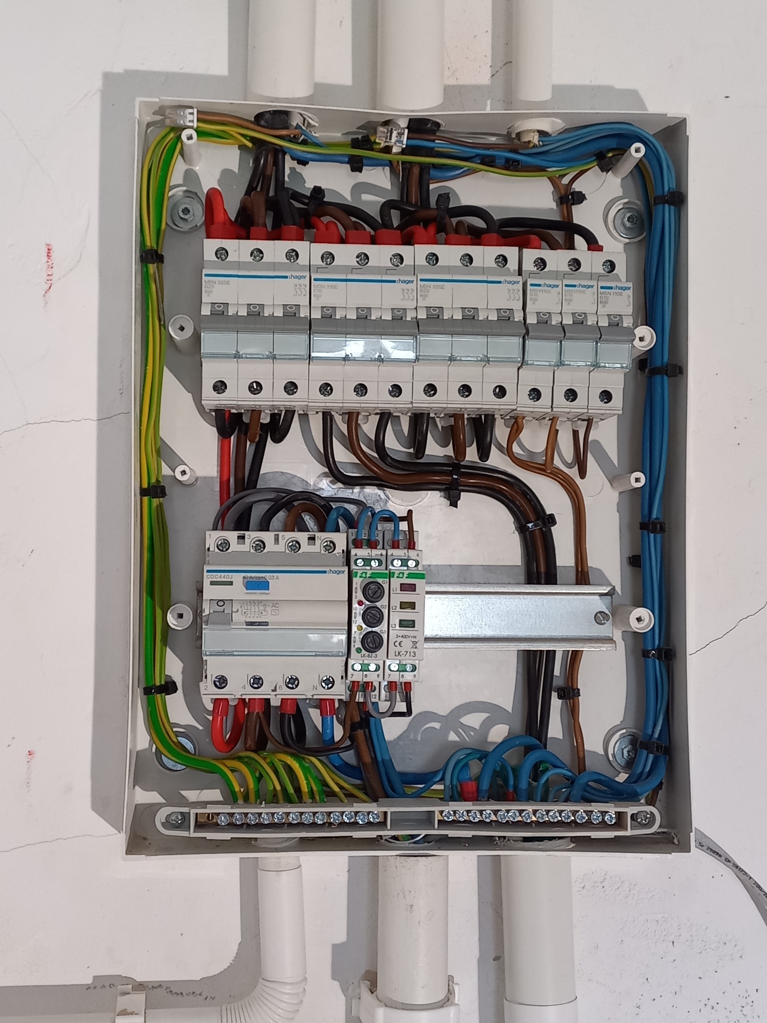 Otwarta skrzynka rozdzielcza z zainstalowanymi bezpiecznikami, przewodami w różnych kolorach (niebieski, żółto-zielony, brązowy, czerwony) i elementami montażowymi, widoczne białe rury instalacyjne...