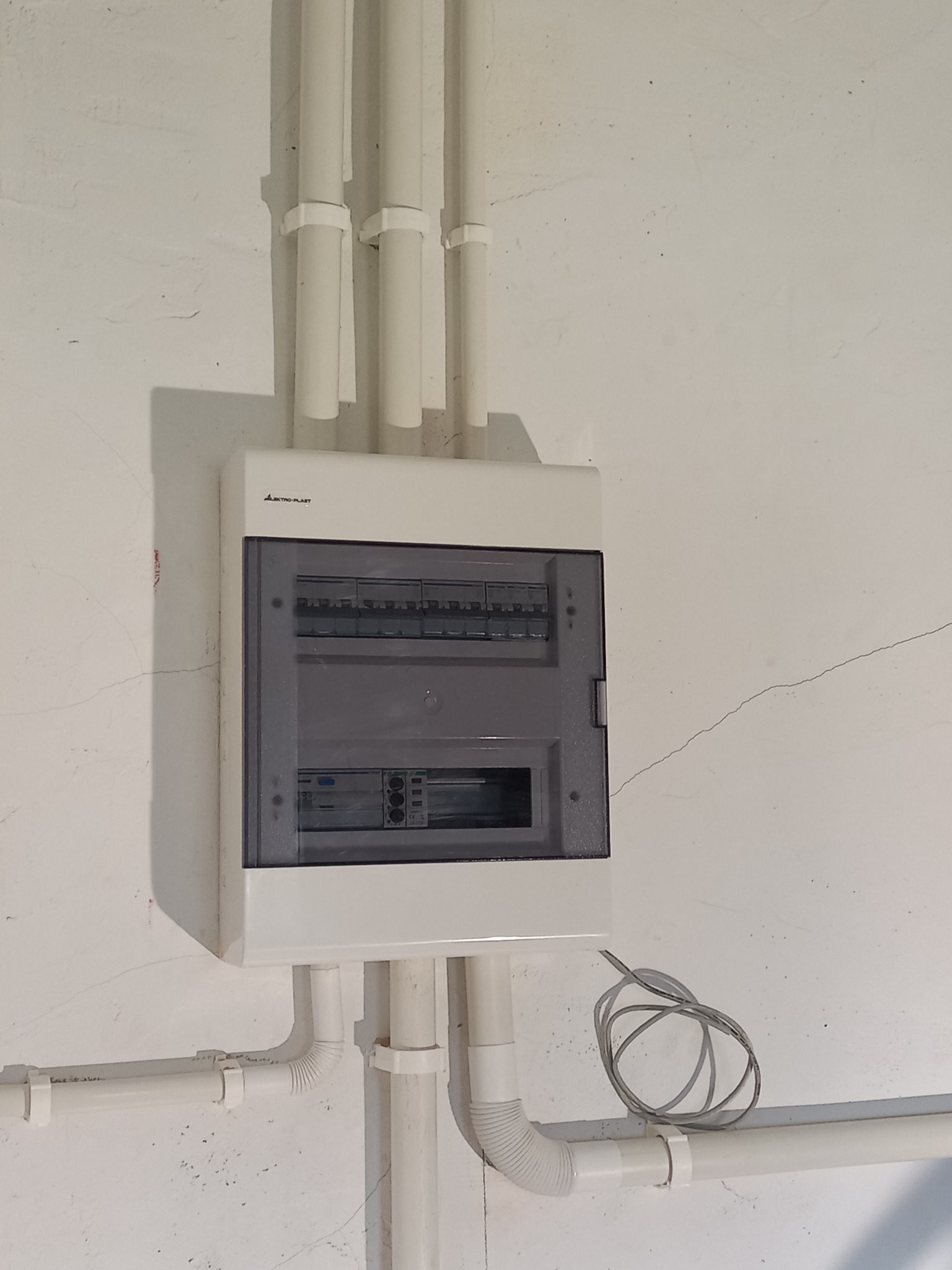 Biała skrzynka rozdzielcza z przezroczystymi drzwiczkami zamontowana na białej, lekko popękanej ścianie, z widocznymi białymi rurami PCV doprowadzającymi przewody.