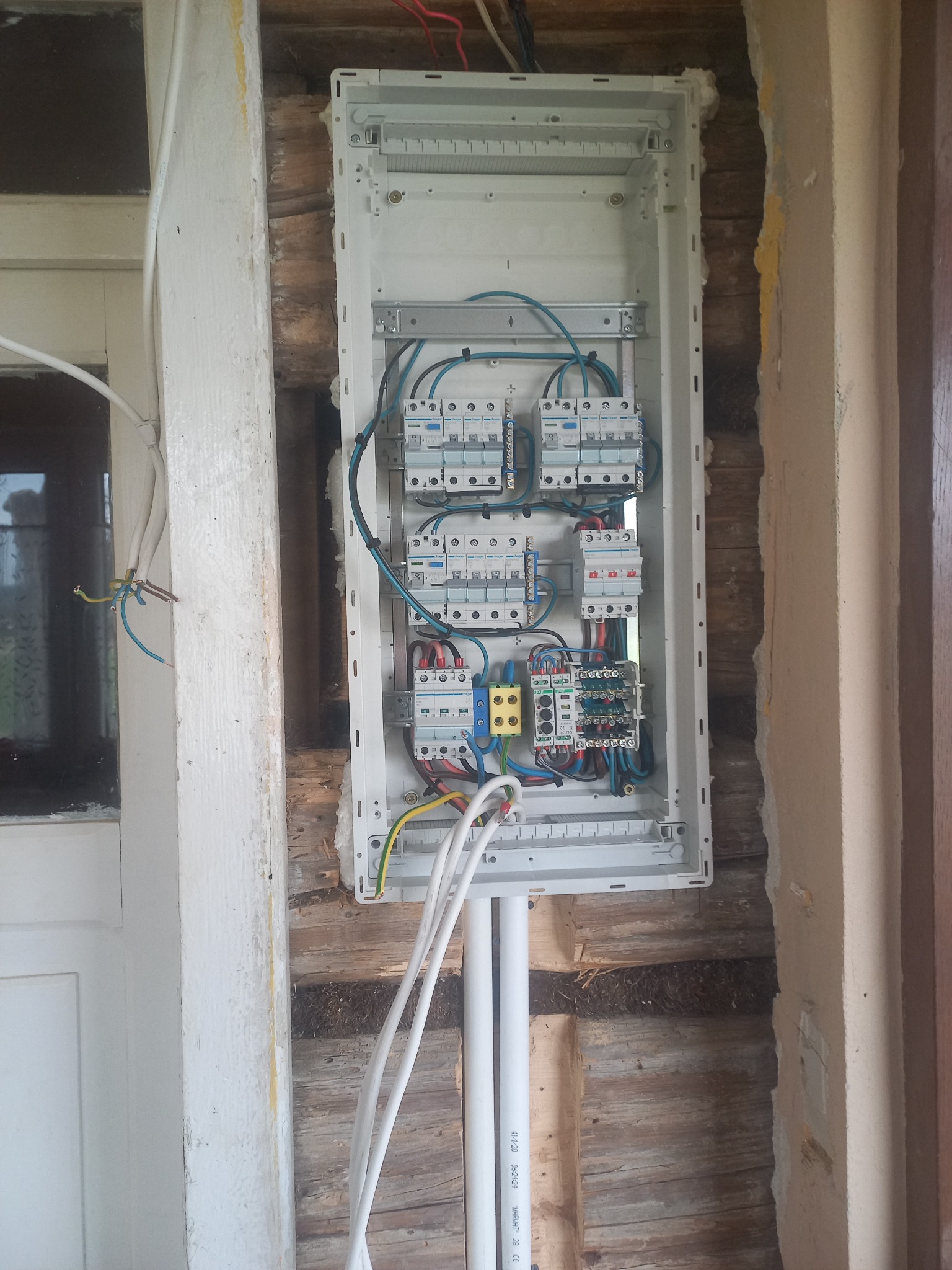 Otwarta skrzynka rozdzielcza z bezpiecznikami i okablowaniem, zamontowana w surowej ścianie z drewnianych bali, widoczne białe rury PCV i wystające przewody elektryczne.