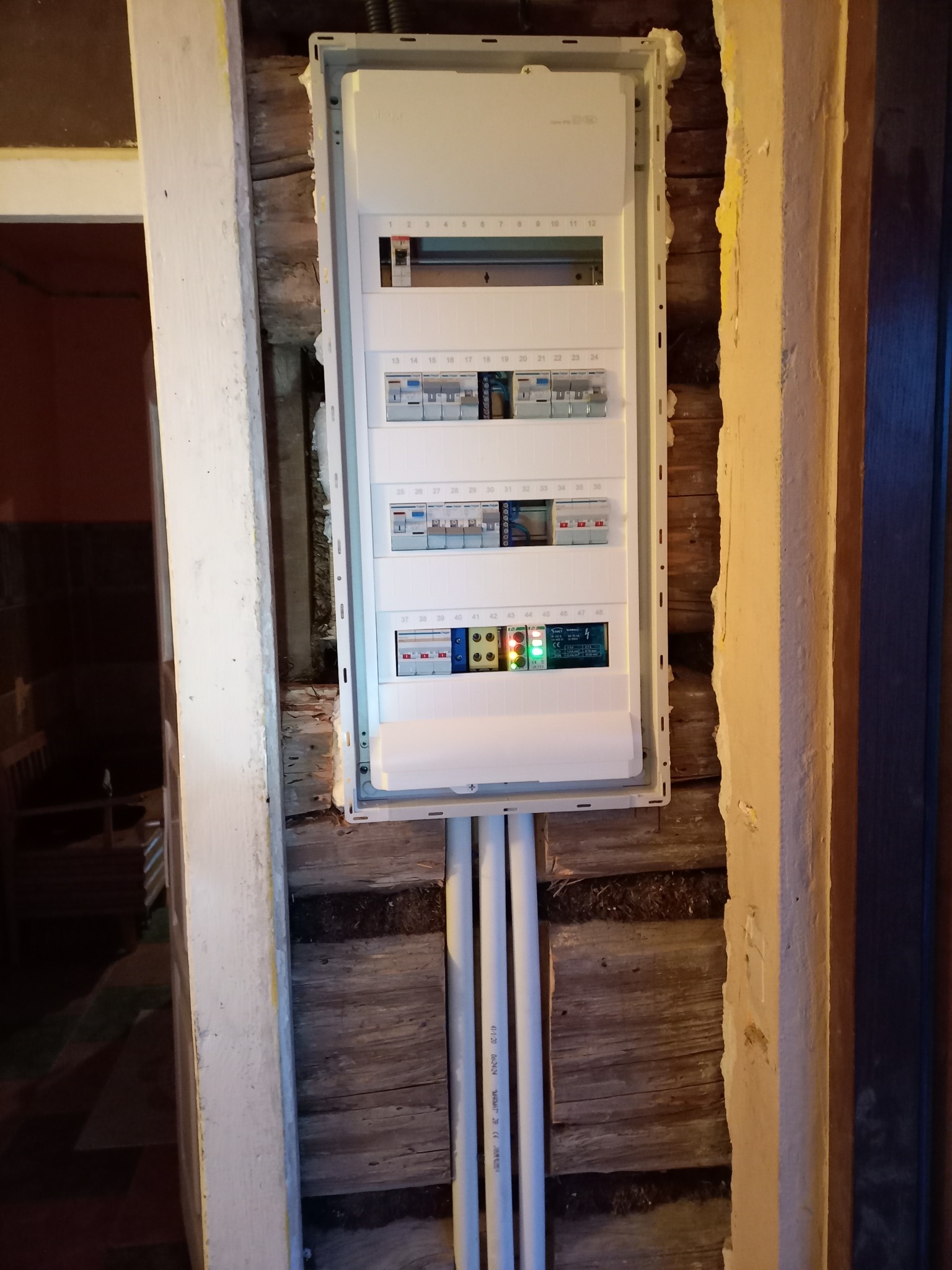 Rozdzielnica elektryczna zamontowana w surowym, drewnianym otworze ściennym z widocznymi kablami w białych rurkach PCV, fragment futryny drzwi po lewej stronie.