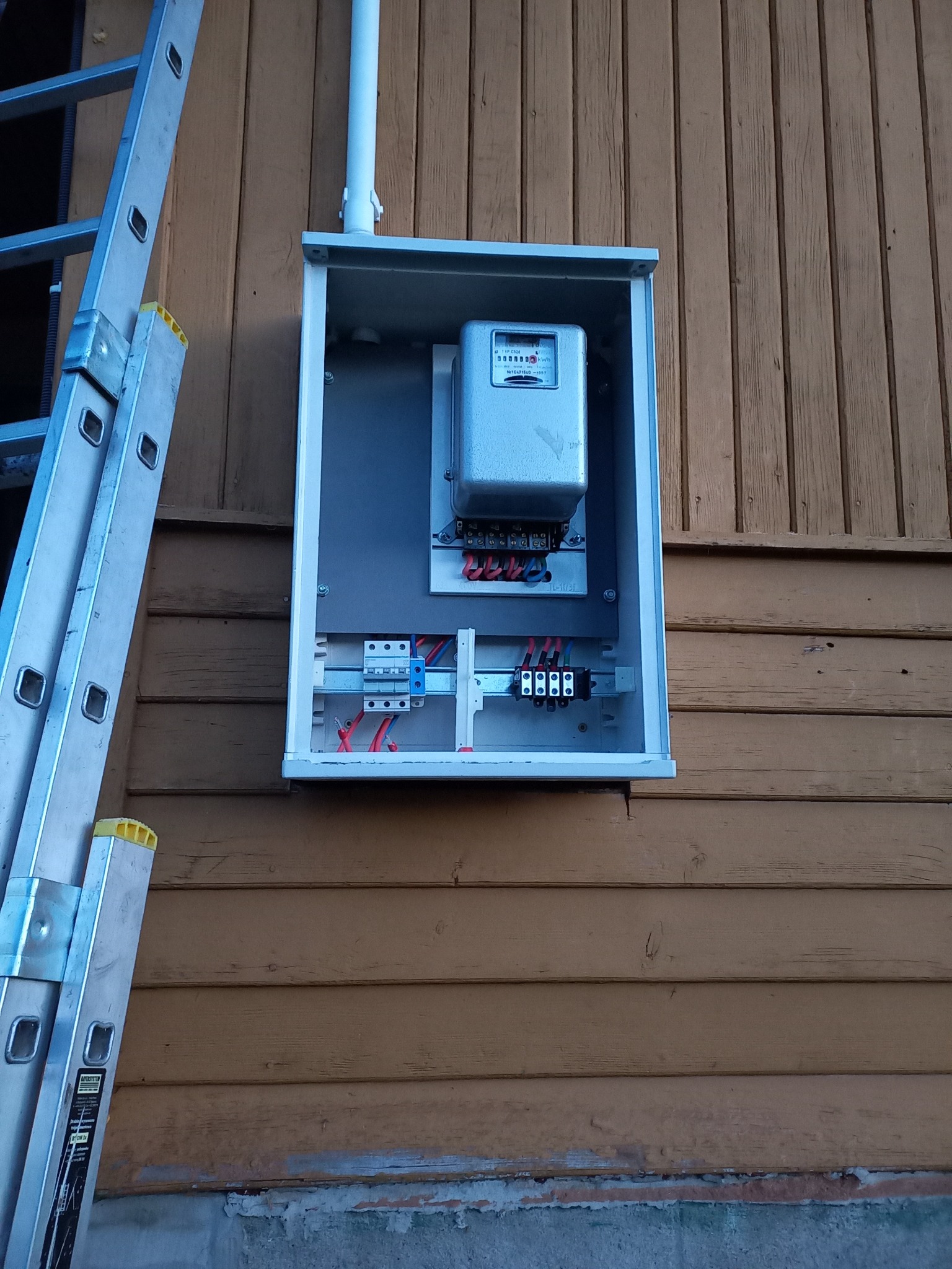 Zewnętrzna skrzynka z licznikiem elektrycznym i przewodami na drewnianej ścianie budynku, obok oparta drabina aluminiowa.