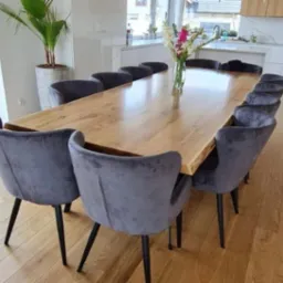 Długi, jasny stół z litego drewna z naturalną krawędzią, otoczony szarymi, welurowymi krzesłami z czarnymi nogami, na drewnianej podłodze; w tle widoczna kuchnia w jasnych kolorach i roślina...