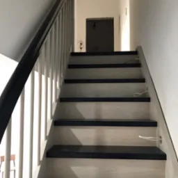 Wewnętrzne schody z drewnianymi stopniami w kolorze czarnym i jasnym drewnem na podstopnicach, z białą balustradą i czarnym poręczą, widoczne przewody elektryczne wystające ze stopni.