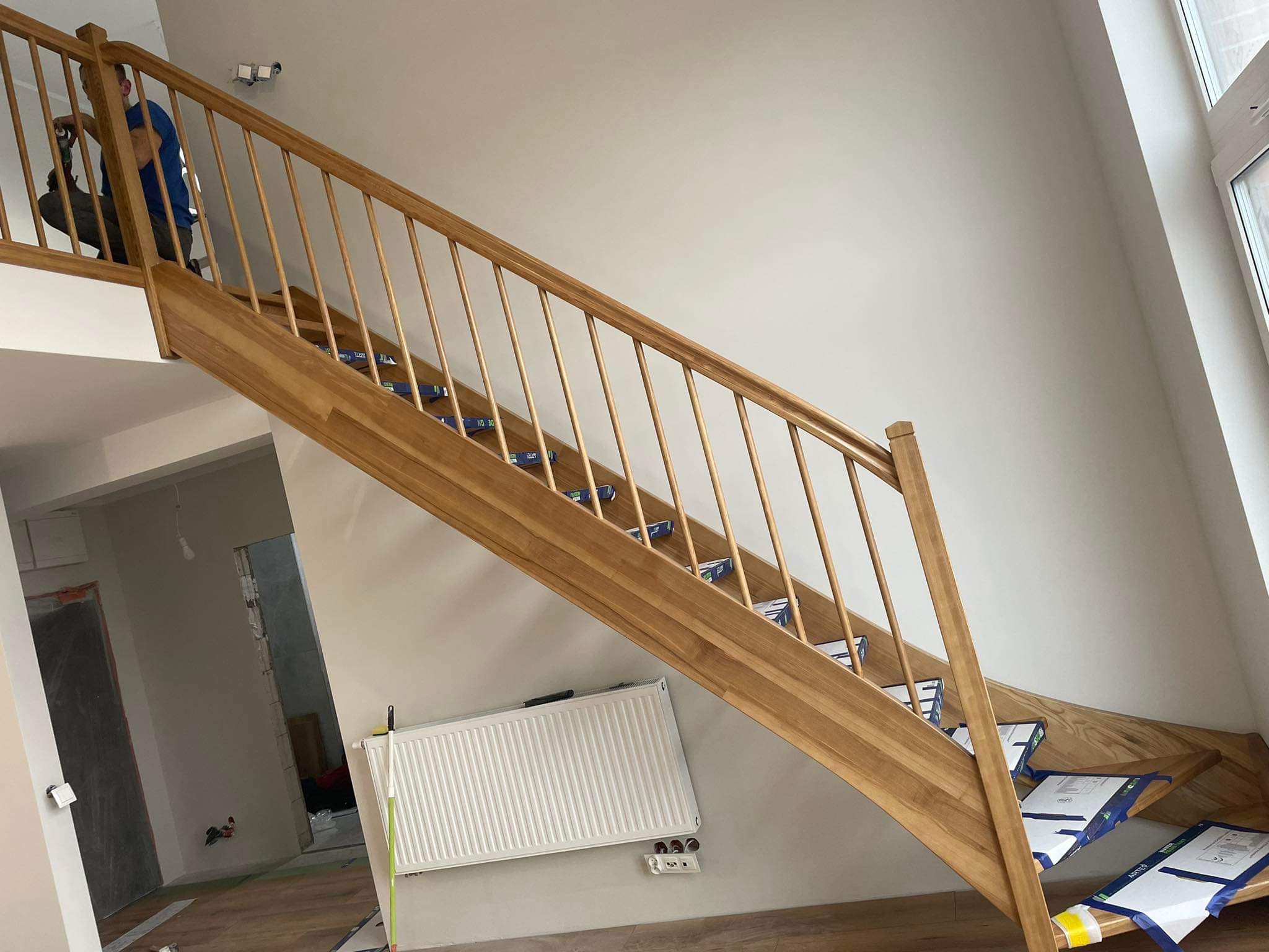 Montaż drewnianych schodów w Dębnie: stolarz wierci w balustradzie, zabezpieczone stopnie, widoczny grzejnik i instalacja elektryczna w tle.