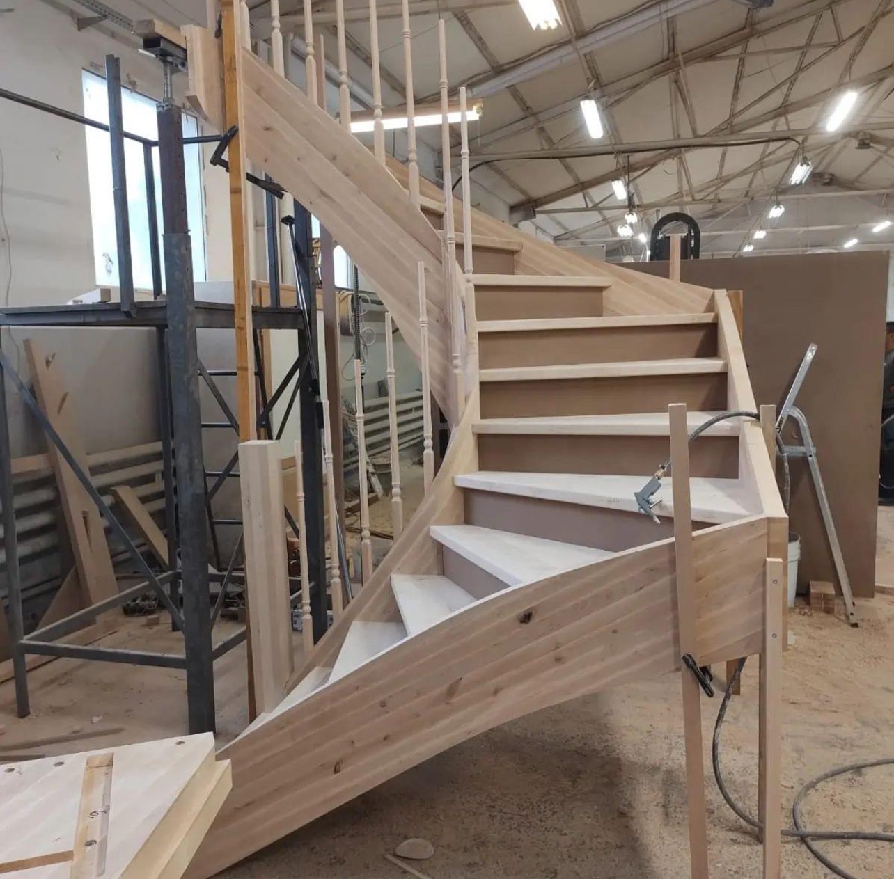 Niedokończona drewniana klatka schodowa z toczonymi balustradami w surowym stanie, widoczne narzędzia i elementy montażowe, hala produkcyjna w tle.