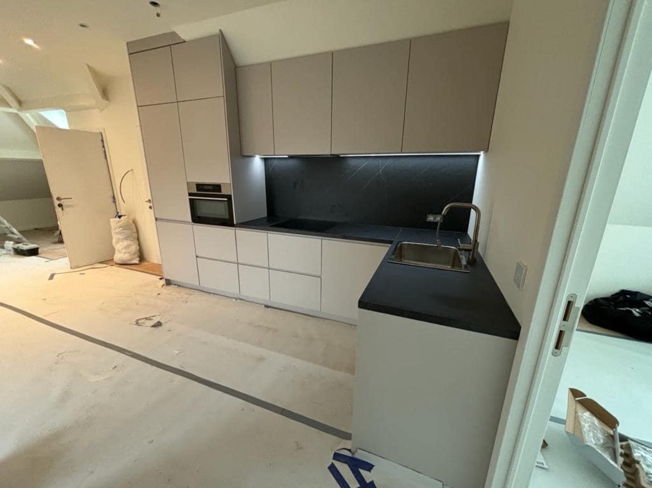 Jasnoszara kuchnia z czarnym blatem i podświetleniem LED pod szafkami wiszącymi w trakcie montażu; widoczne niedokończone podłogi i otwarte drzwi.