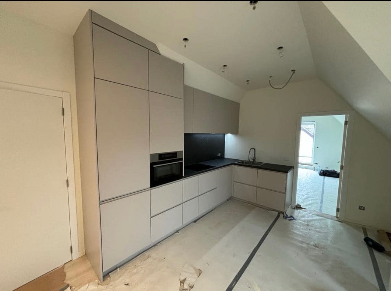 Jasna kuchnia z zabudową pod skosem dachu, czarny blat i backsplash, widoczne niedokończone wykończenie podłogi.