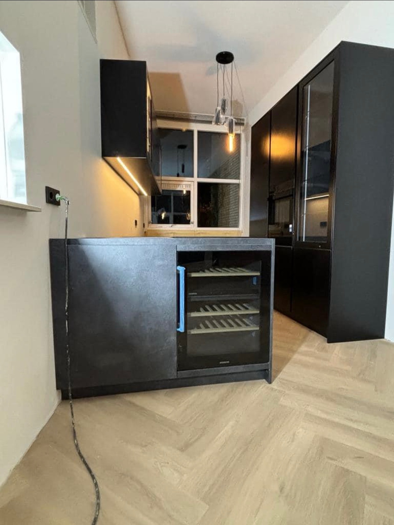 Nowoczesna kuchnia w odcieniach szarości i czerni z drewnianą podłogą w jodełkę; widoczna zabudowa kuchenna z wbudowaną chłodziarką do wina i oświetleniem LED pod szafkami wiszącymi.