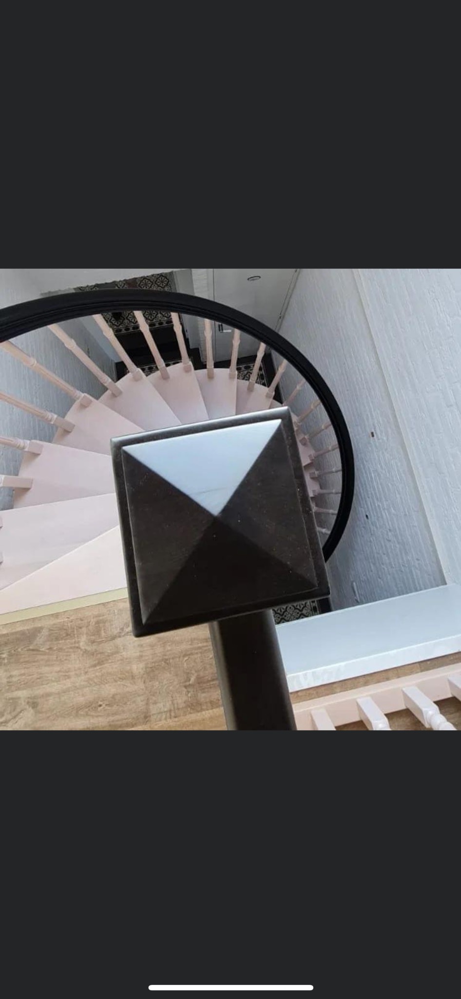 Spiralne schody z drewnianymi stopniami w odcieniach różu, czarną balustradą i kwadratowym, ciemnym słupkiem, widok z góry.