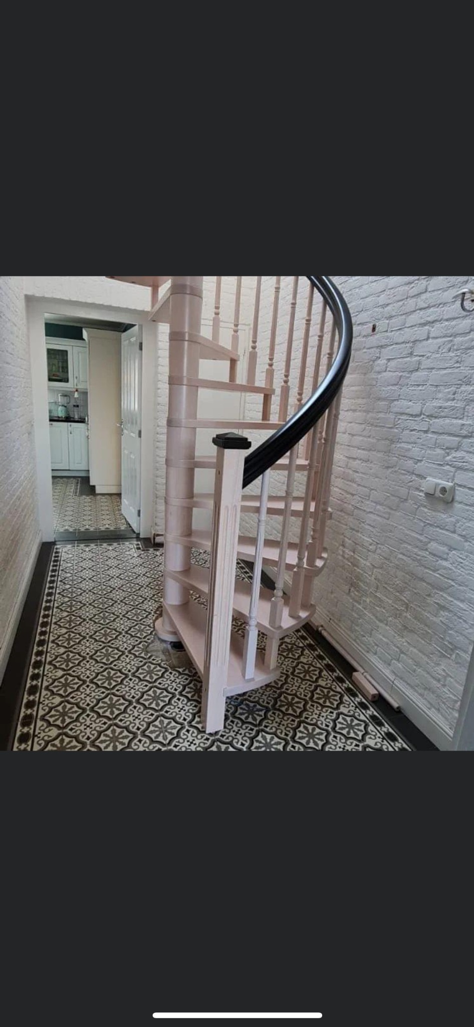 Jasnoróżowe spiralne schody z czarną poręczą w białym korytarzu z wzorzystą podłogą prowadzą do kuchni w tle.