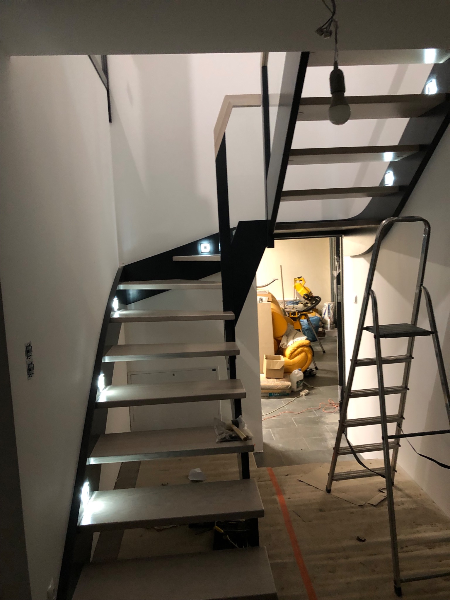 Nowoczesne schody z podświetleniem LED na stopniach, czarne barierki, widoczna drabina i narzędzia w tle, sugerujące trwający montaż.