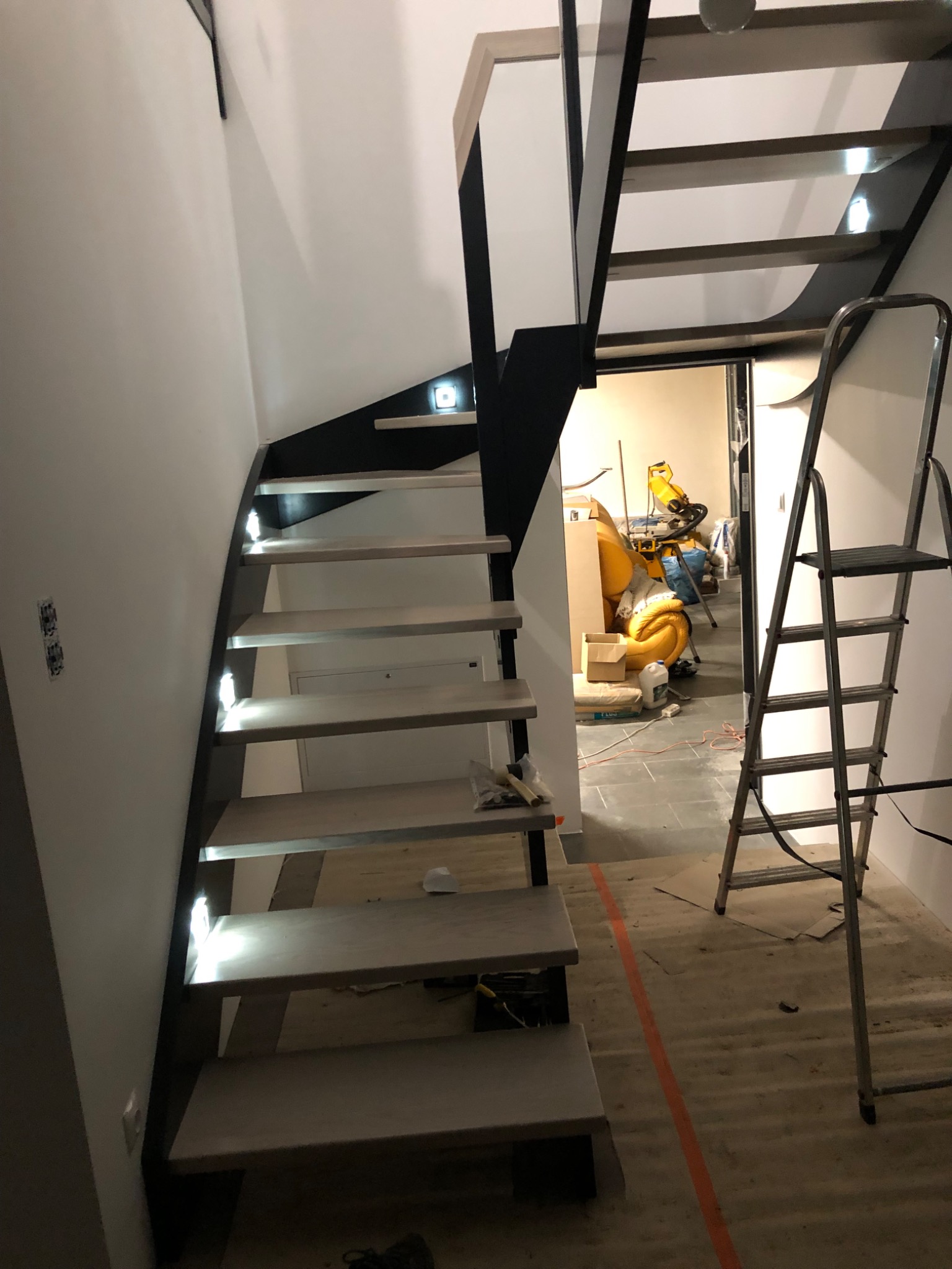 Schody wewnętrzne z drewnianymi stopniami i czarną metalową konstrukcją, podświetlane diodami LED, w trakcie montażu, widoczna drabina i narzędzia w tle.