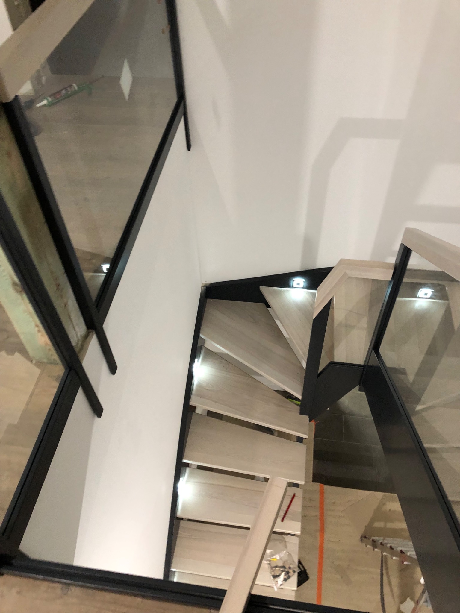 Nowoczesne schody z jasnego drewna z podświetleniem LED w stopniach i szklaną balustradą z czarnymi ramami, widok z góry w trakcie montażu, narzędzia widoczne w tle.