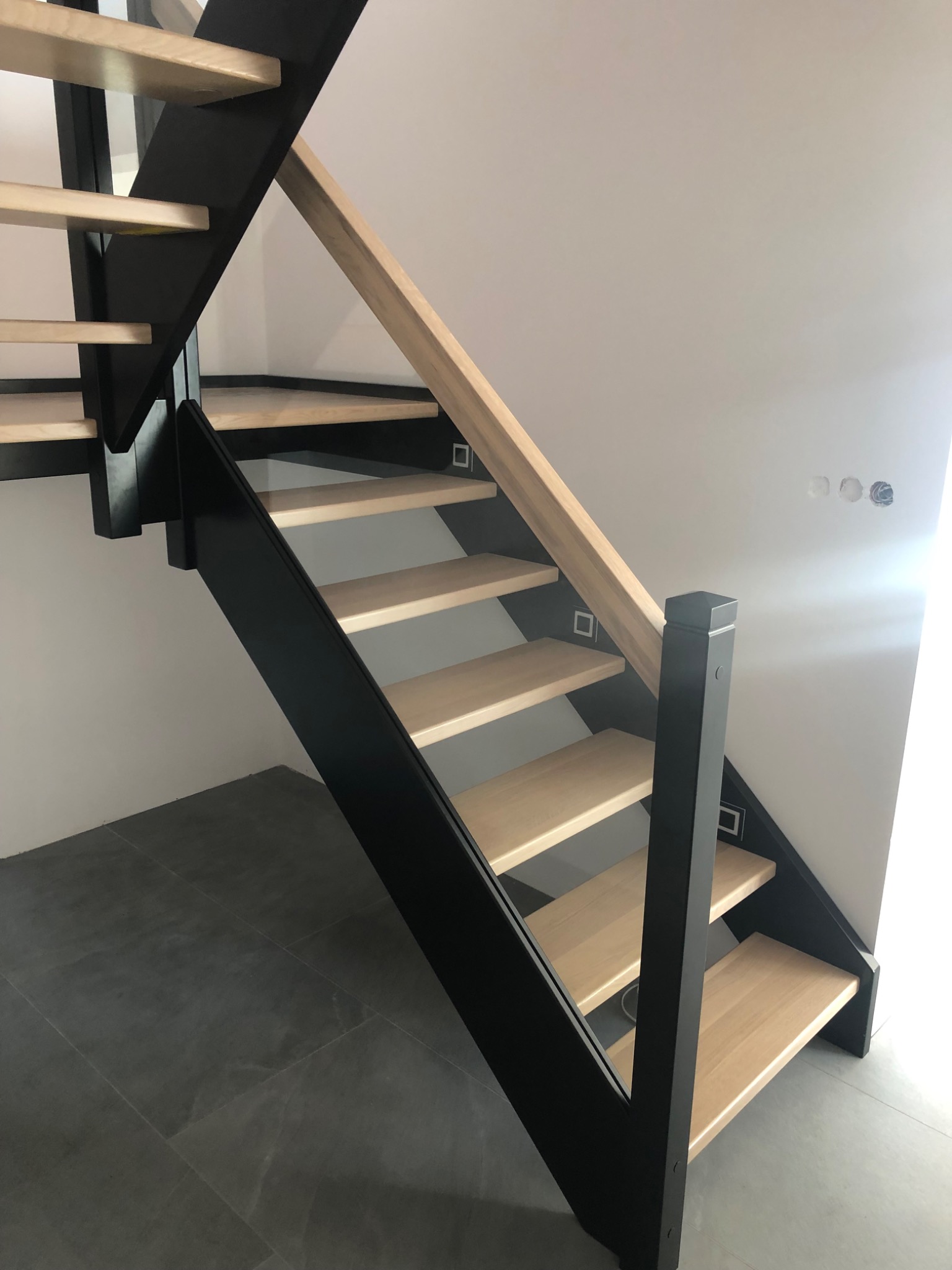 Nowoczesne schody z jasnego drewna z czarną konstrukcją, widok z dołu na tle białej ściany z widocznymi puszkami elektrycznymi.