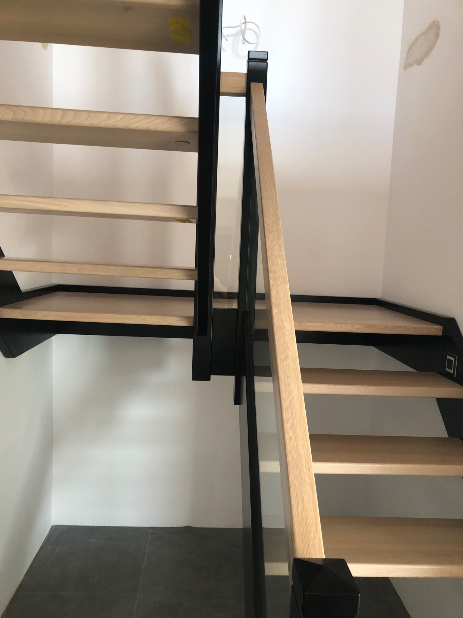 Nowoczesne schody z jasnego drewna i czarnymi elementami konstrukcyjnymi, szklana balustrada, widok z dołu na górę, minimalistyczne wykończenie wnętrza.