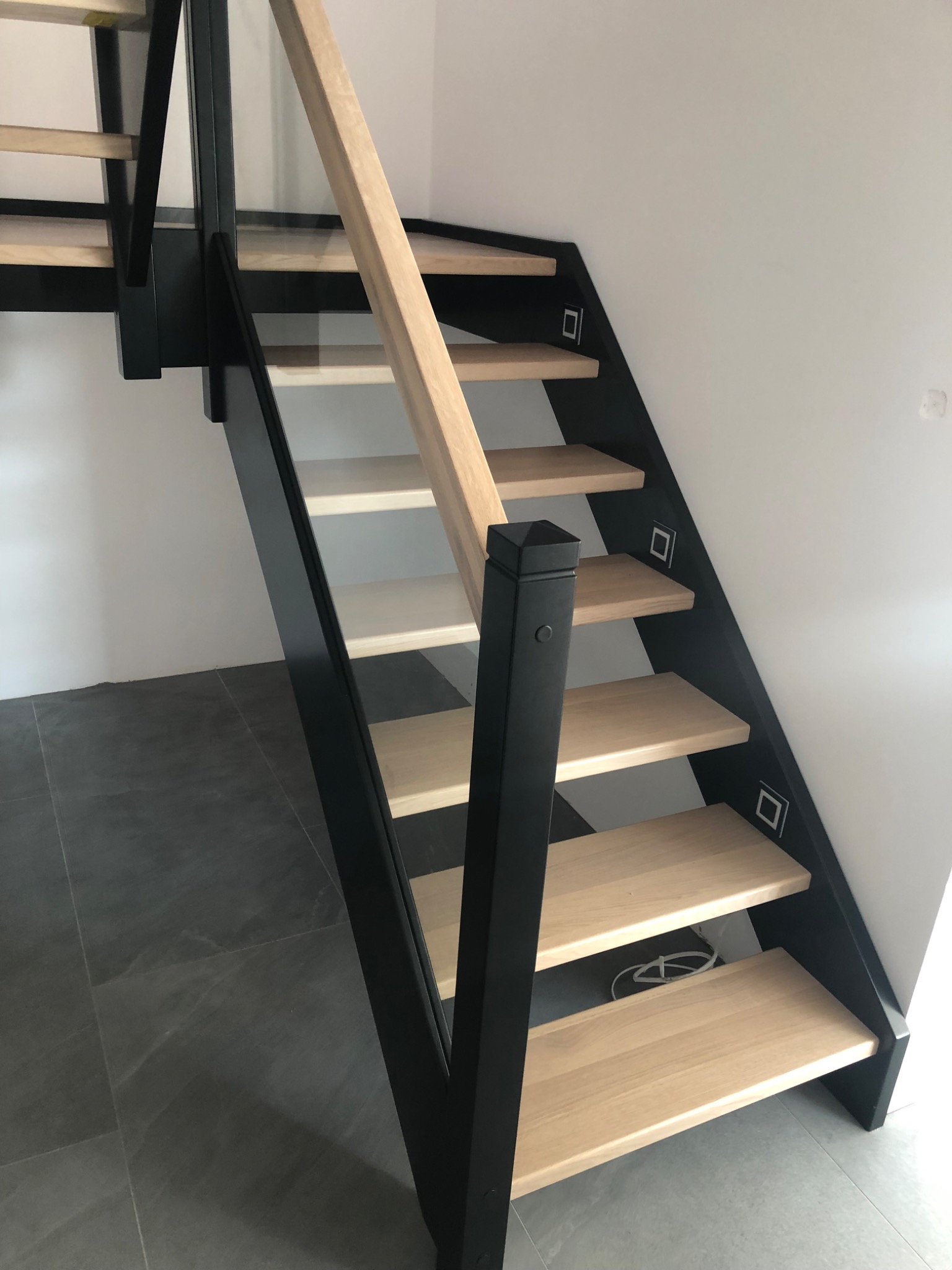 Nowoczesne schody drewniane z czarną balustradą i szklanymi panelami, oświetlenie LED wbudowane w stopnice, widok z dołu na szarej podłodze.