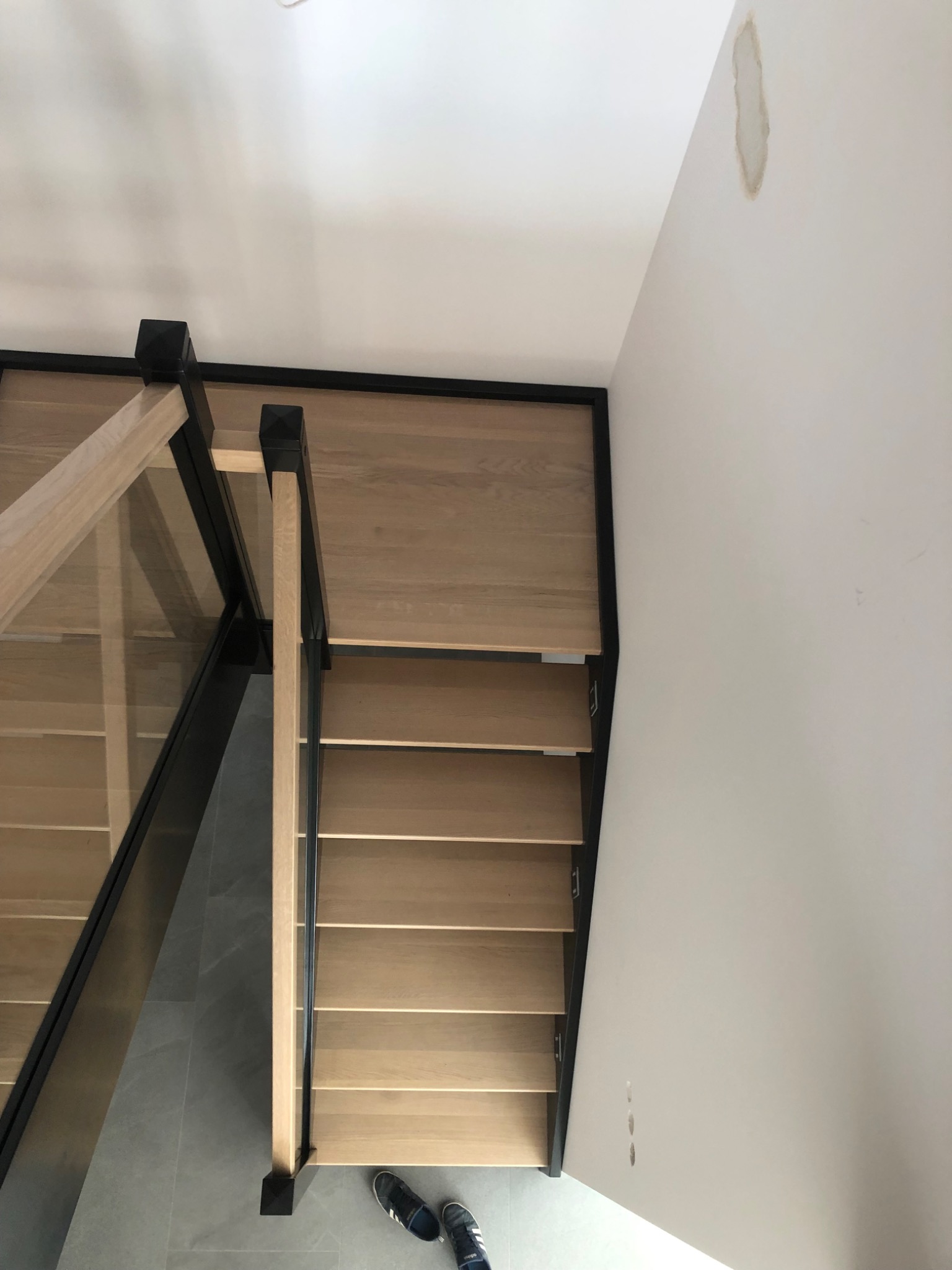Nowoczesne schody z drewnianymi stopniami, czarną balustradą i szklanym wypełnieniem, widok z góry, fragment ściany z plamą.