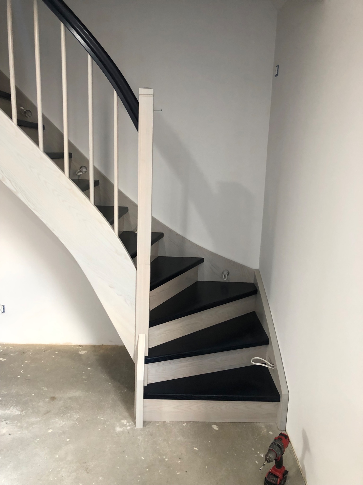 Biało-czarne schody z drewnianymi balustradami na tle surowej ściany, w dolnym rogu wiertarka akumulatorowa, widoczne okablowanie oświetlenia.