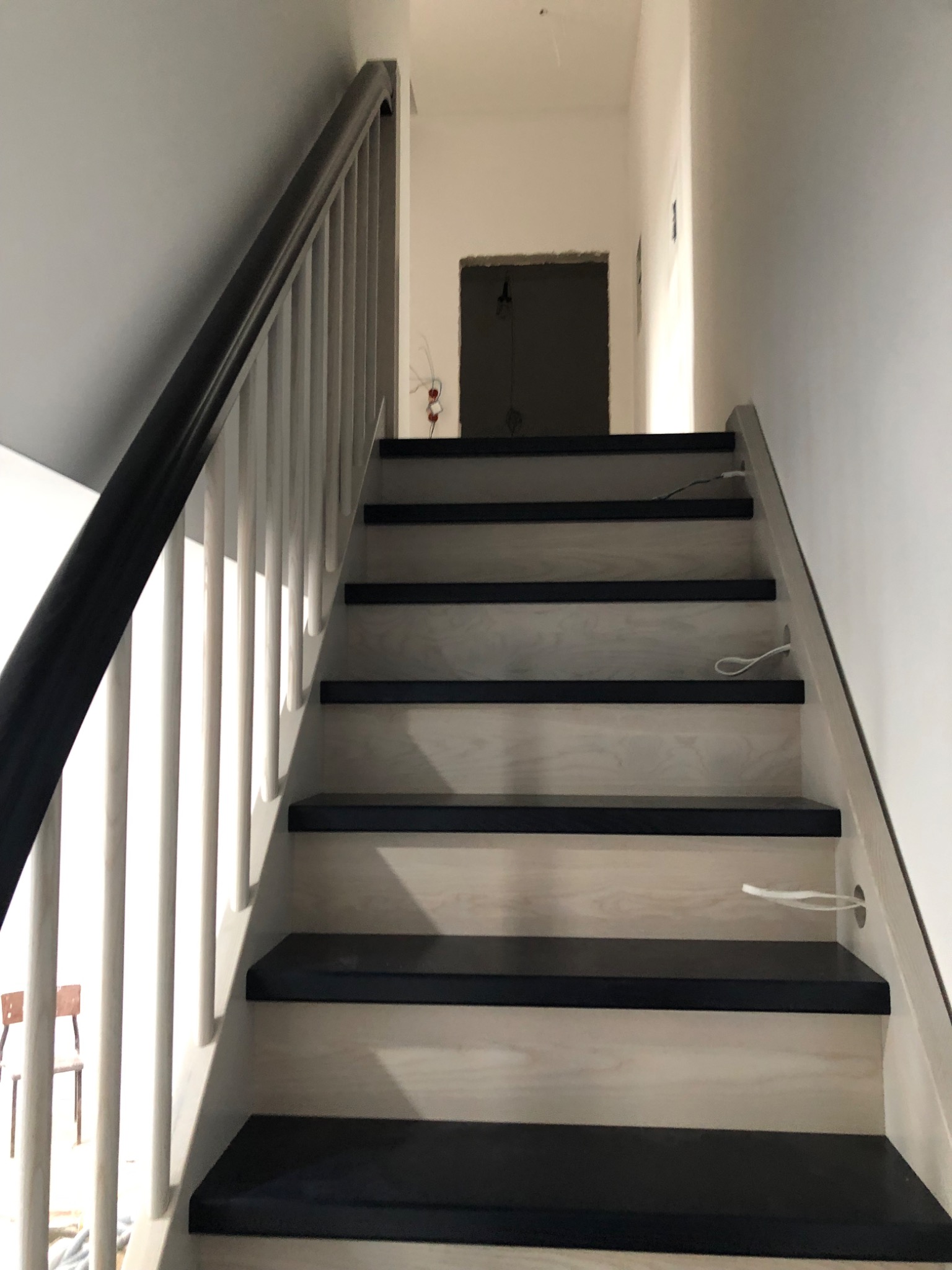 Wewnętrzne schody z drewnianymi stopniami w kolorze czarnym i jasnym drewnem na podstopnicach, z białą balustradą i czarnym poręczą, widoczne przewody elektryczne wystające ze stopni.