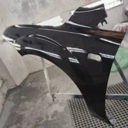 Czarny, błyszczący błotnik samochodowy zawieszony na metalowym stojaku w lakierni, widoczne odbicia światła na lakierowanej powierzchni, w tle kask motocyklowy i inne elementy karoserii.