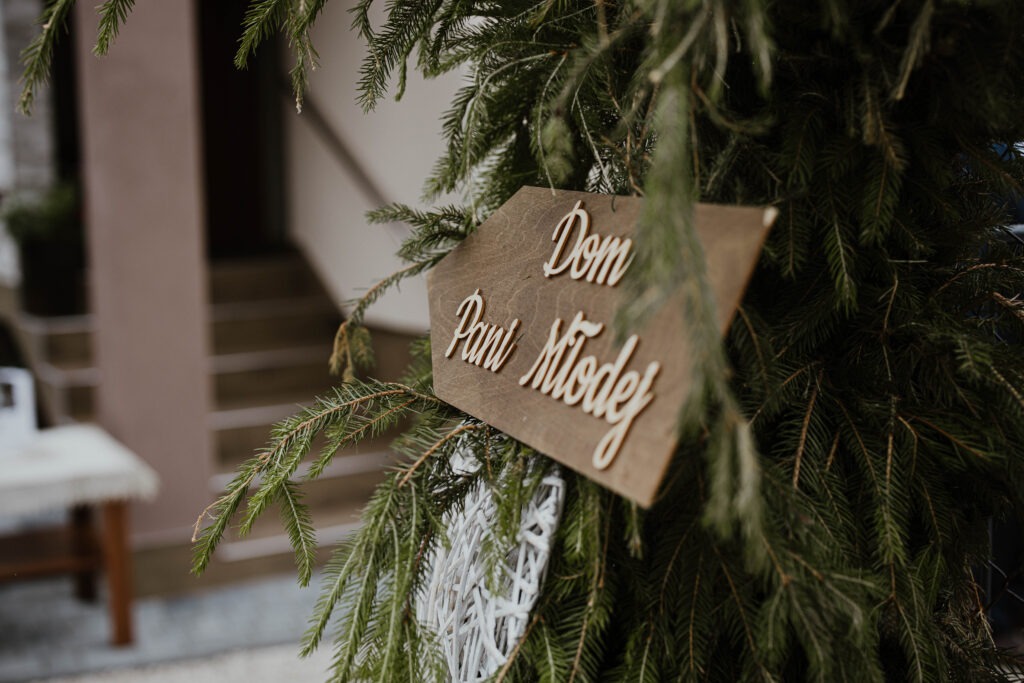 Drewniany drogowskaz z napisem 'Dom Pani Młodej' zawieszony na gałęziach choinki, w tle schody i budynek.
