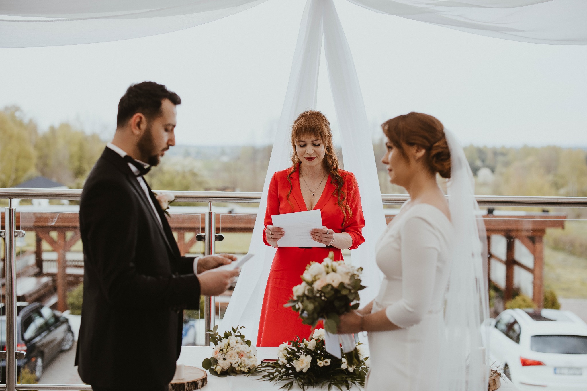 Para młoda podczas ceremonii ślubnej na balkonie, panna młoda trzyma bukiet, w tle las i samochody.