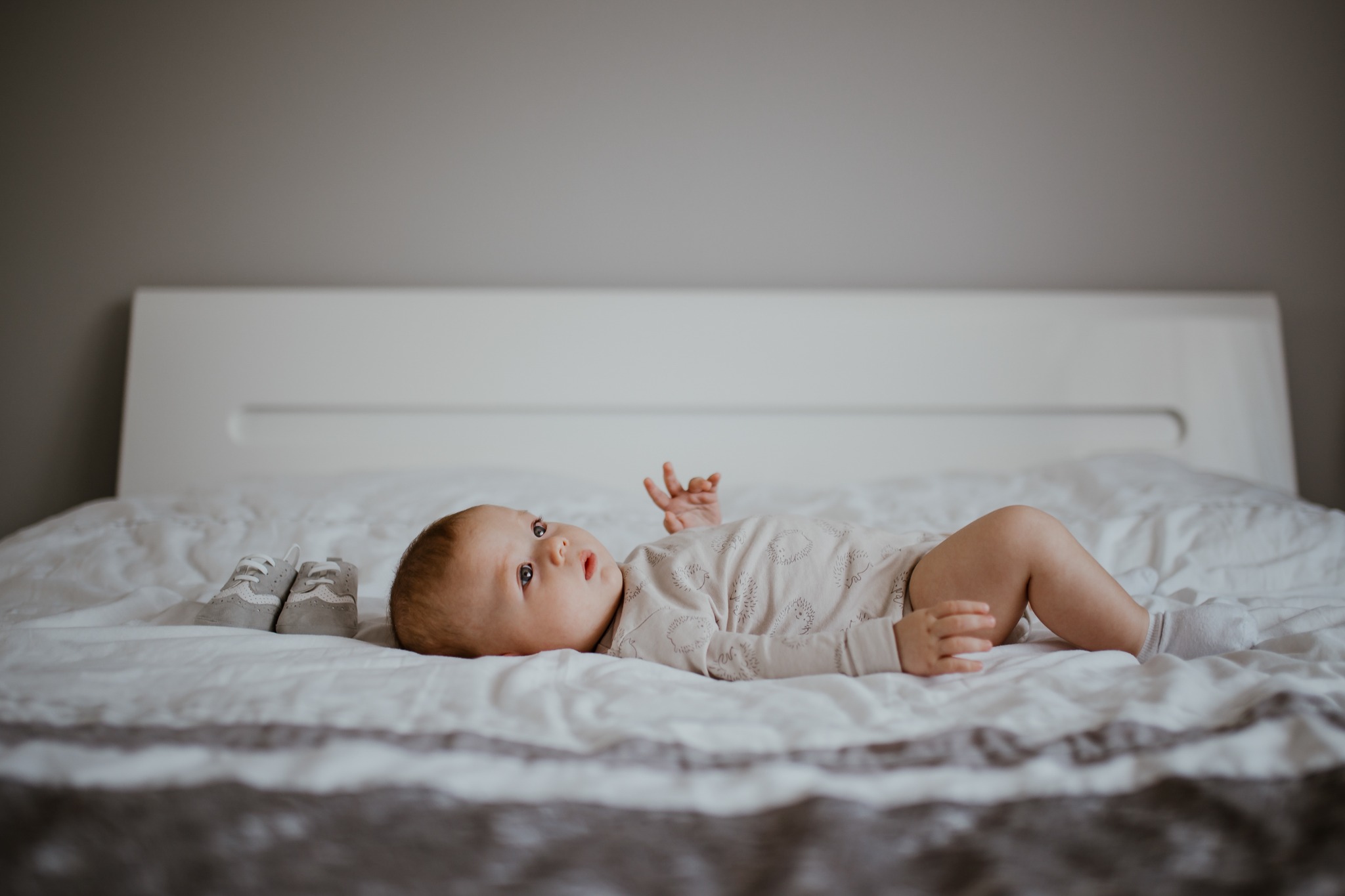Niemowlę leży na łóżku w beżowym body z motywem jeży, obok leżą malutkie szare buciki.