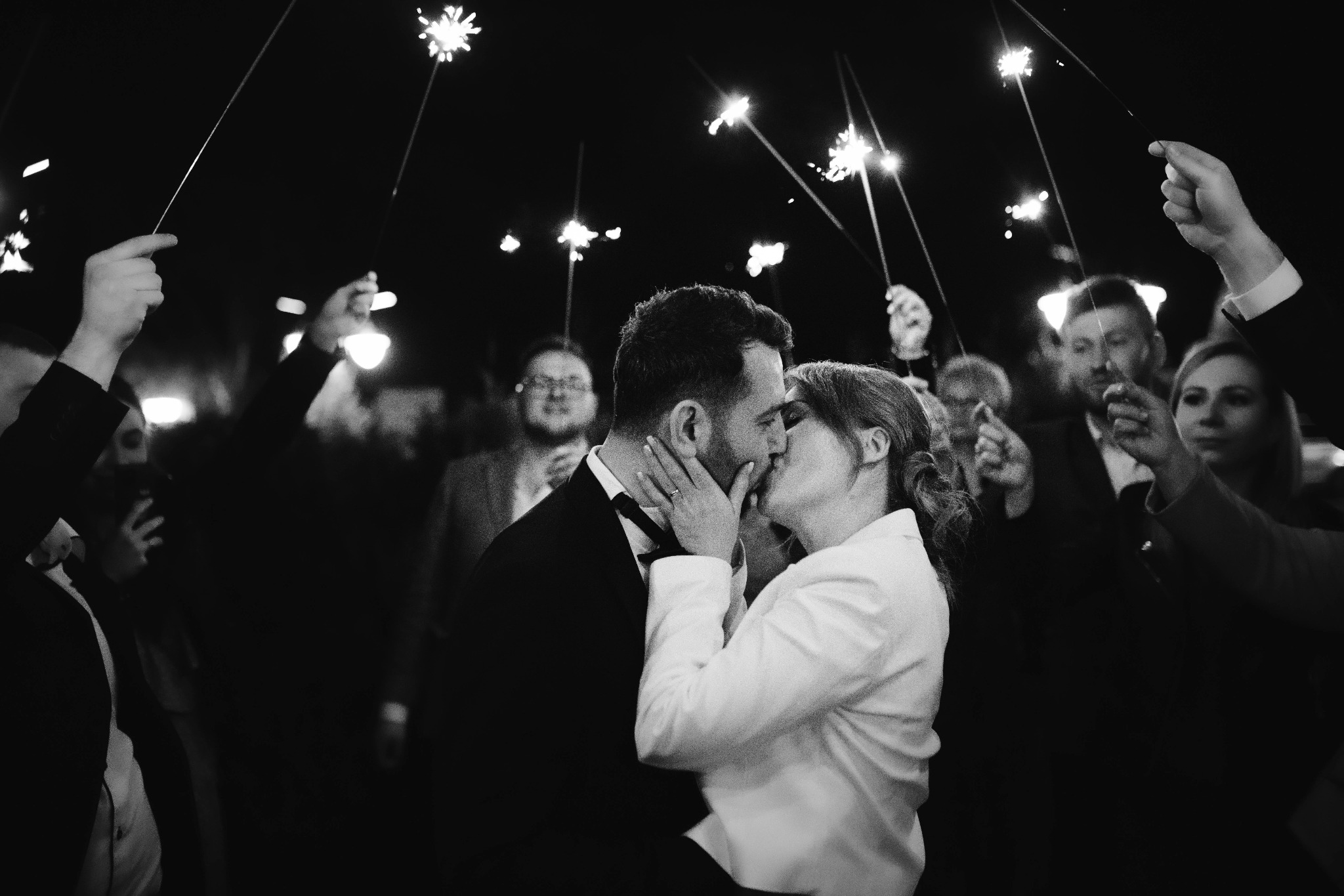 Czarno-białe zdjęcie całującej się pary młodej otoczonej przez gości z zimnymi ogniami, z widocznym pierścionkiem zaręczynowym na dłoni panny młodej.